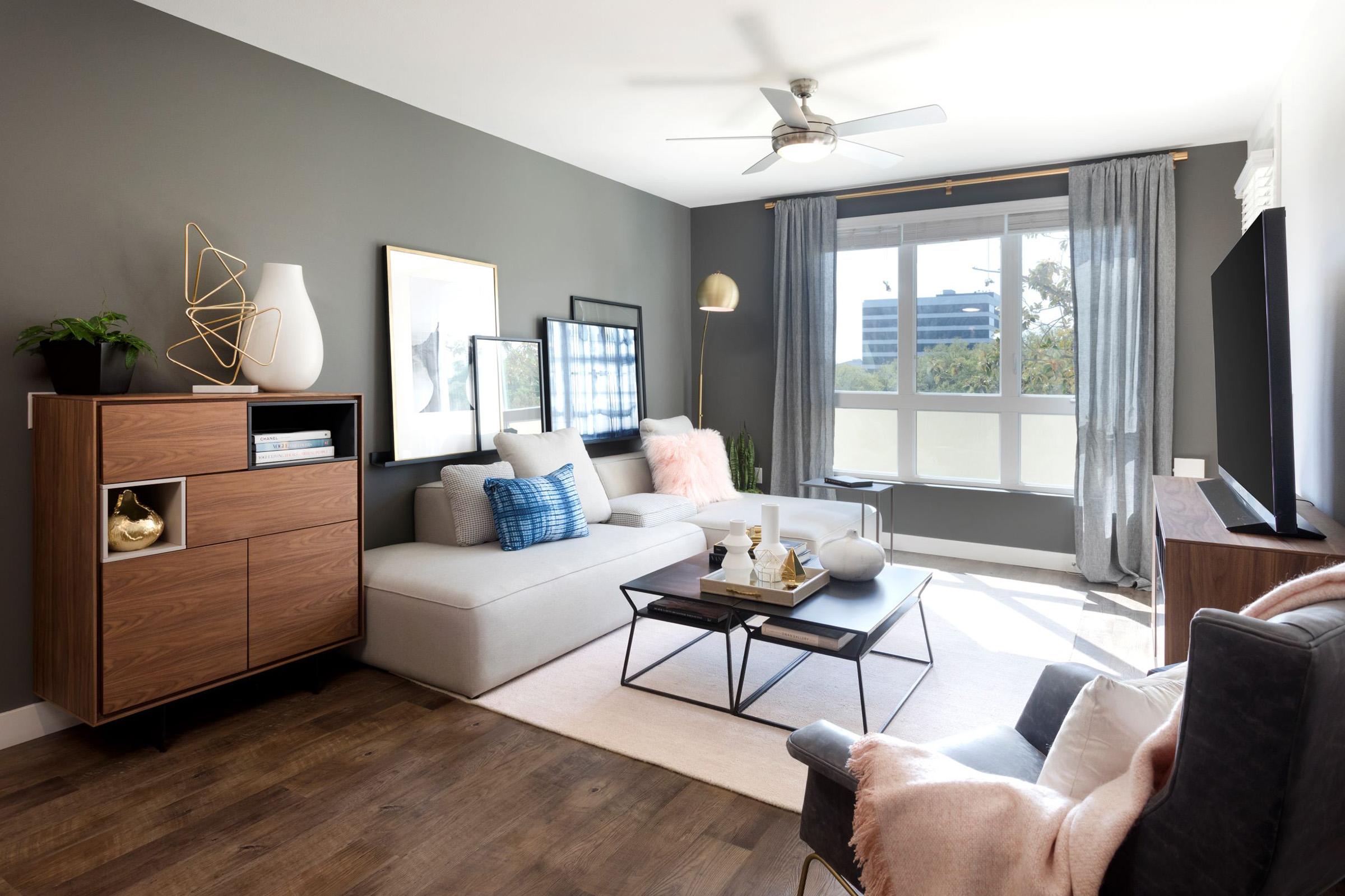 A modern living room with a light gray sectional sofa and a dark wooden cabinet. There’s a coffee table with decorative items in the center, and large windows with sheer curtains allowing natural light. A ceiling fan is visible, along with plants and artwork on the walls, creating a cozy atmosphere.