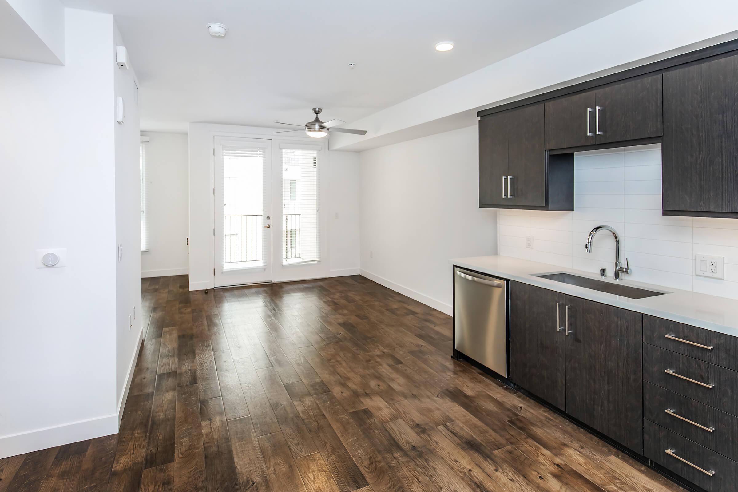 Modern kitchen with dark wood cabinets and stainless steel appliances, including a dishwasher. Light-colored countertop and backsplash. Open living space with large windows and a door leading to a balcony. Hardwood flooring throughout. Ceiling fan in the room. Bright and airy atmosphere.