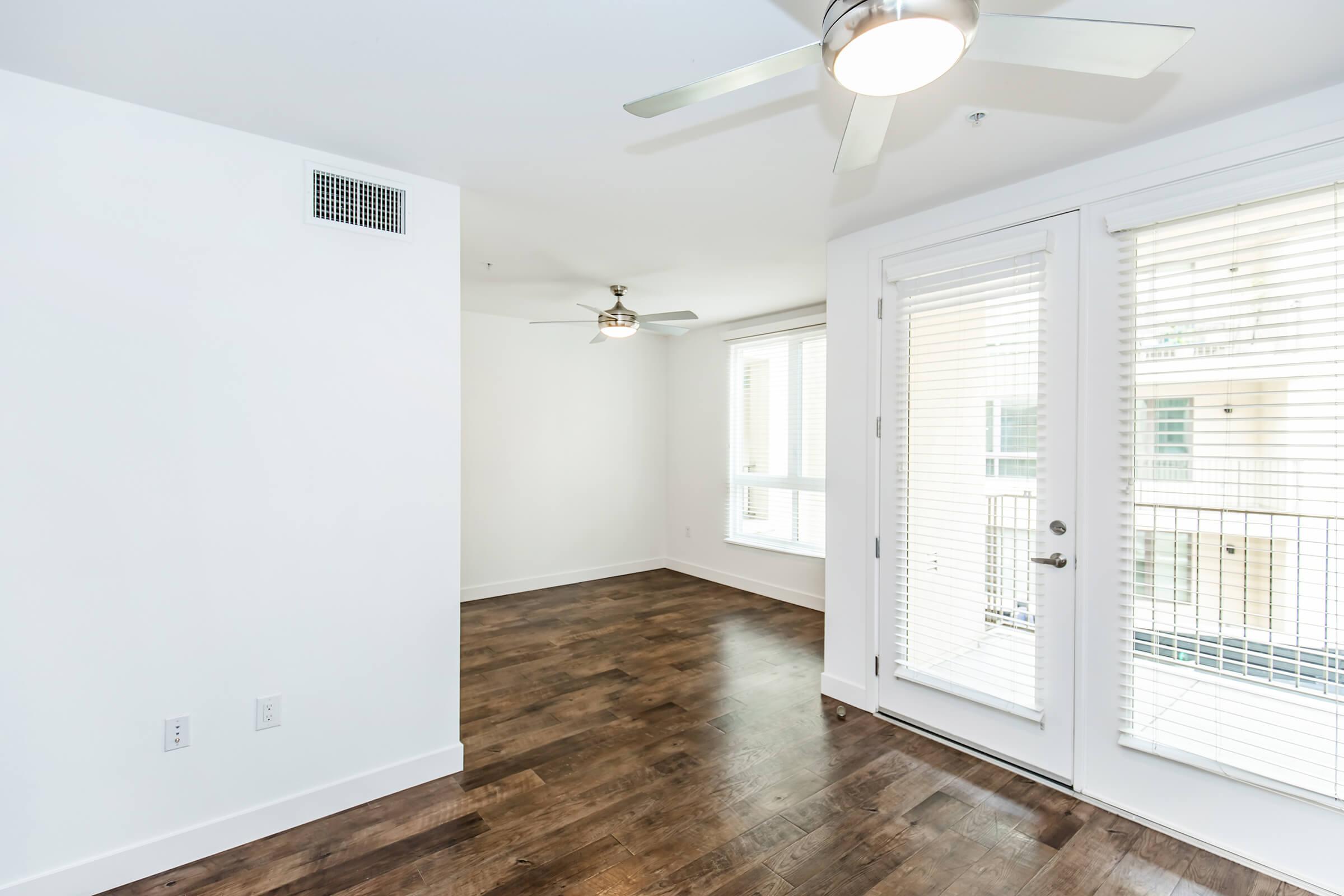 A well-lit, modern living space featuring light-colored walls and hardwood floors. There are two ceiling fans and large windows with blinds. A door leads to a small balcony, and the layout is open, providing a spacious feel.