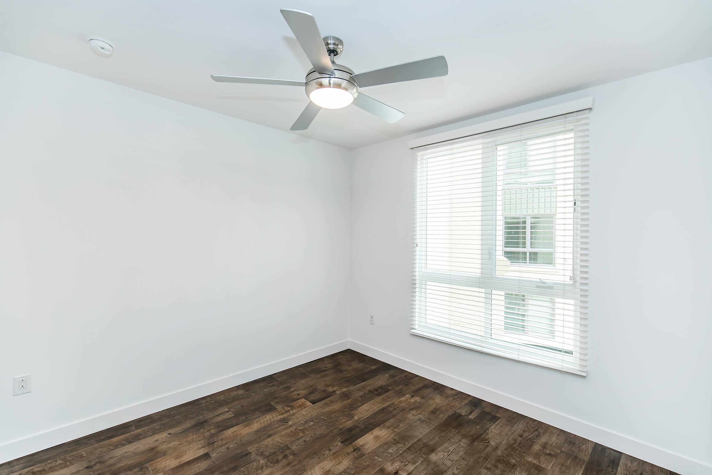 A spacious, minimally decorated room featuring a ceiling fan, large window with blinds, and brown wooden flooring. The walls are painted white, creating a bright and airy atmosphere.