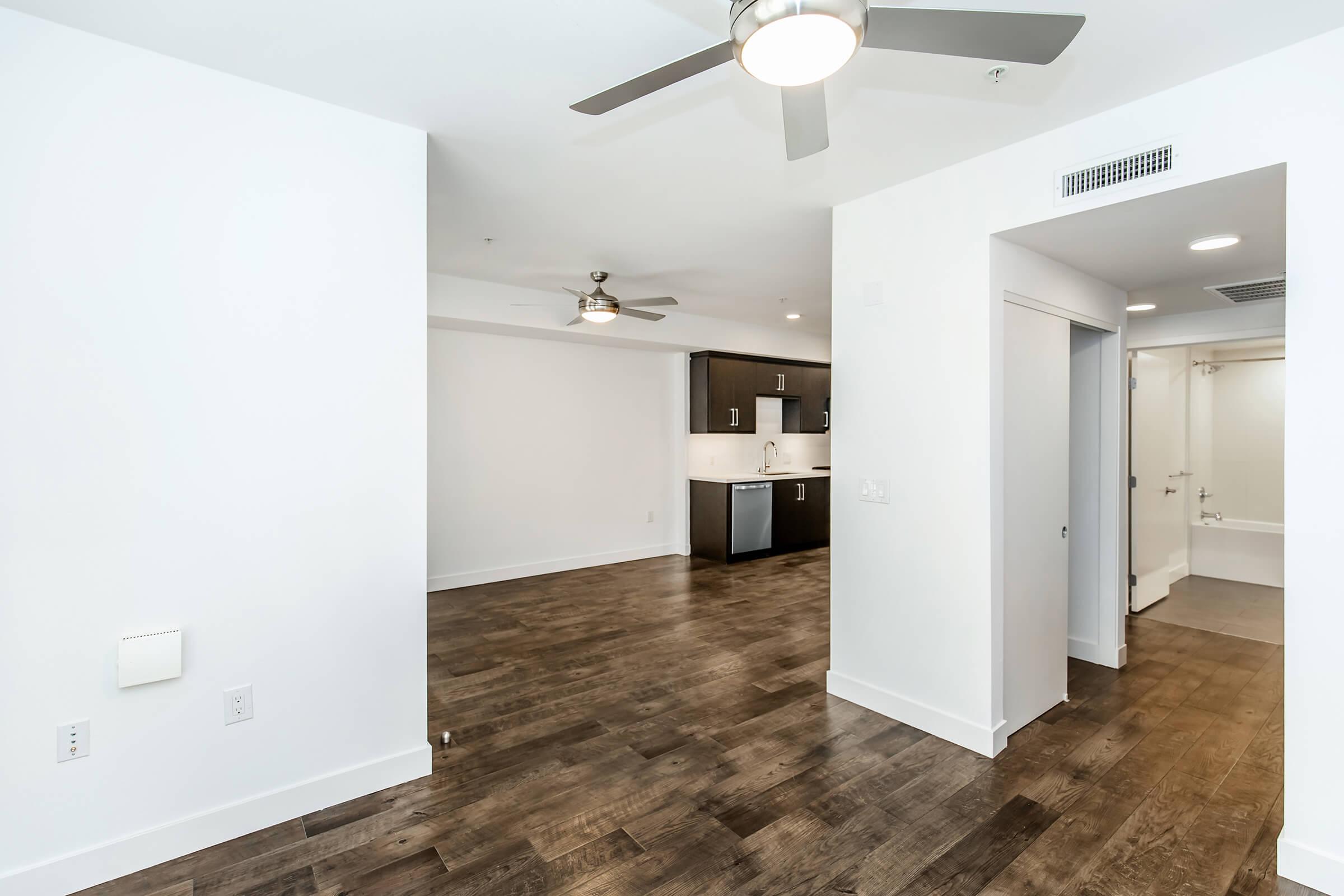 Bright and modern open-concept living space with hardwood floors, featuring a kitchen in the background with dark cabinetry and stainless steel appliances. Two ceiling fans provide airflow, and glass lighting fixtures add brightness. A doorway on the right leads to a bathroom or utility area.