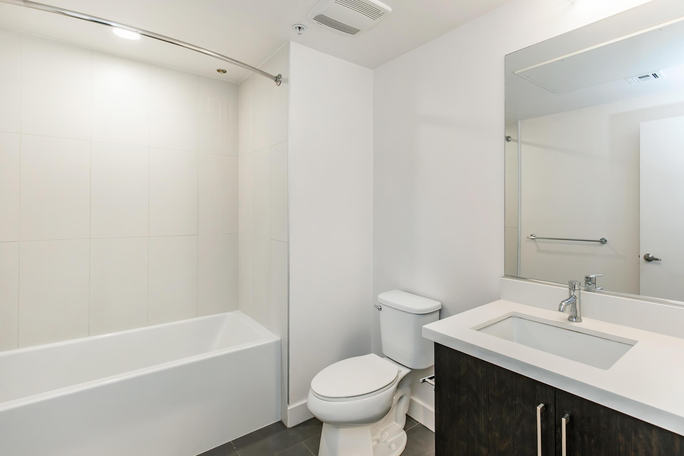 A modern bathroom featuring a white bathtub with a shower, a toilet, and a dark wood vanity with a white countertop and sink. The space is brightly lit with neutral-colored walls and minimal decor, creating a clean and spacious appearance.