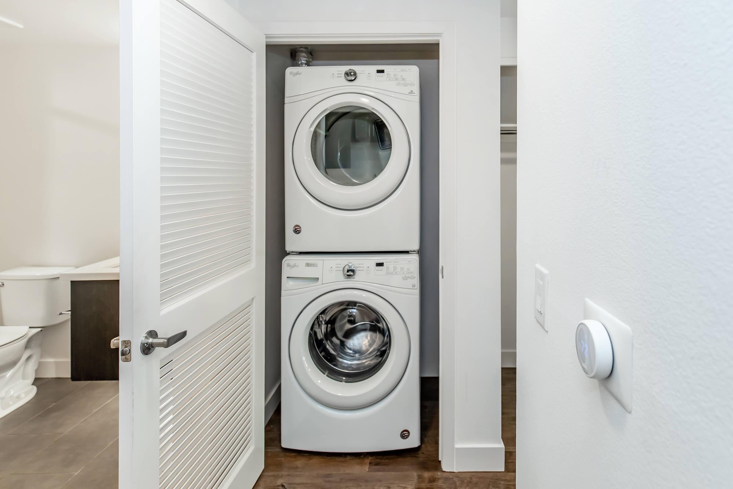 A stacked washer and dryer in a narrow closet space, with white doors and light-colored walls. The room features wood flooring, and a glimpse of a bathroom with a toilet can be seen in the background.