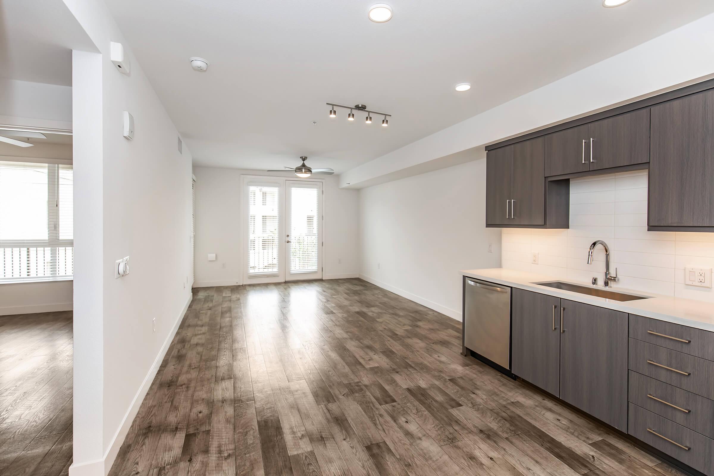 Modern open-concept kitchen and living area featuring hardwood floors, stainless steel appliances, and contemporary cabinets. Large windows provide natural light, with sliding glass doors leading to a balcony. Neutral walls enhance the spacious feel.