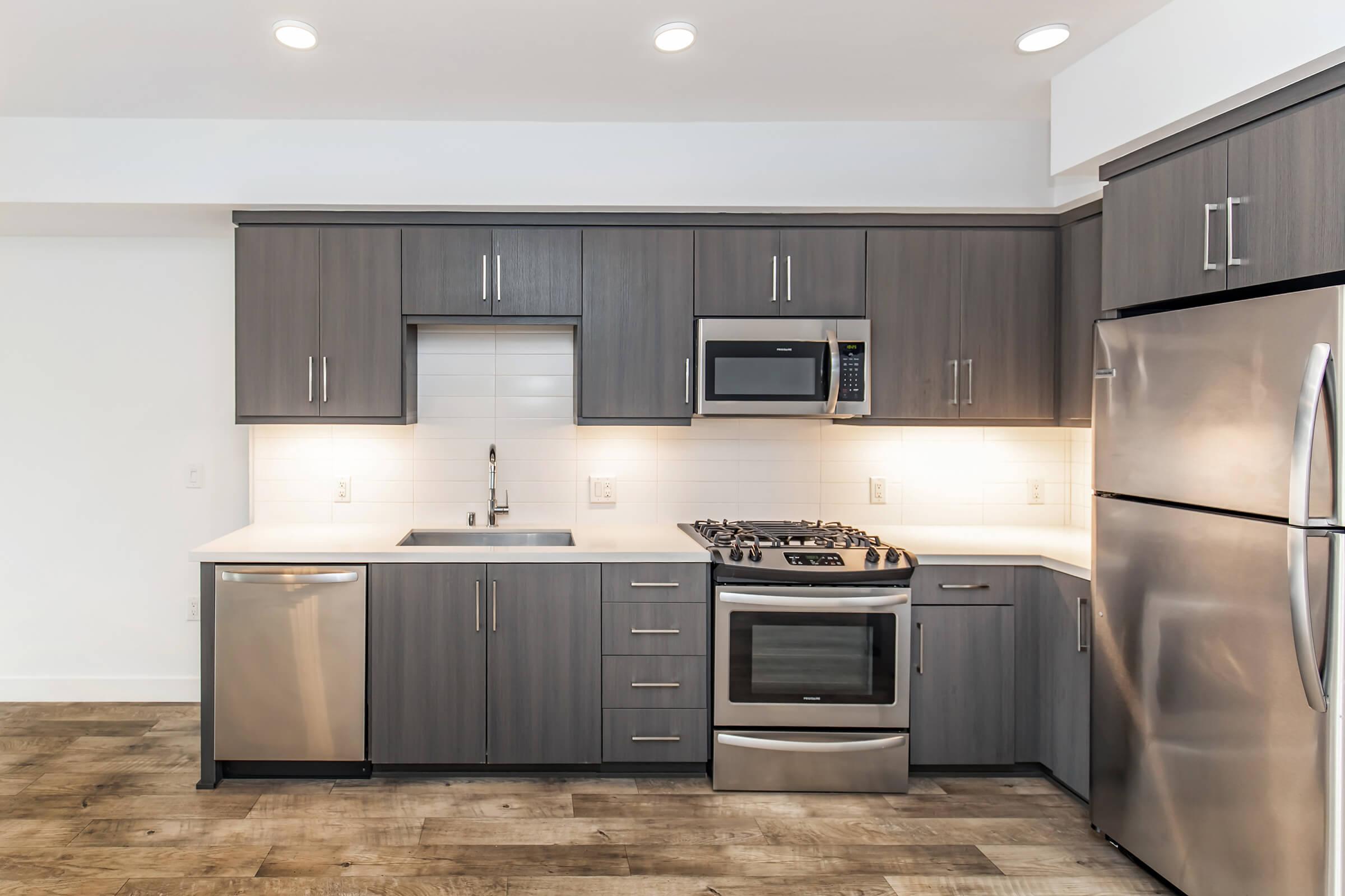 Modern kitchen with gray cabinetry, stainless steel appliances, including a gas stove, microwave, and refrigerator. White tile backsplash and countertop create a clean aesthetic, complemented by recessed lighting and wood-like flooring. The sink is positioned under the window for natural light.