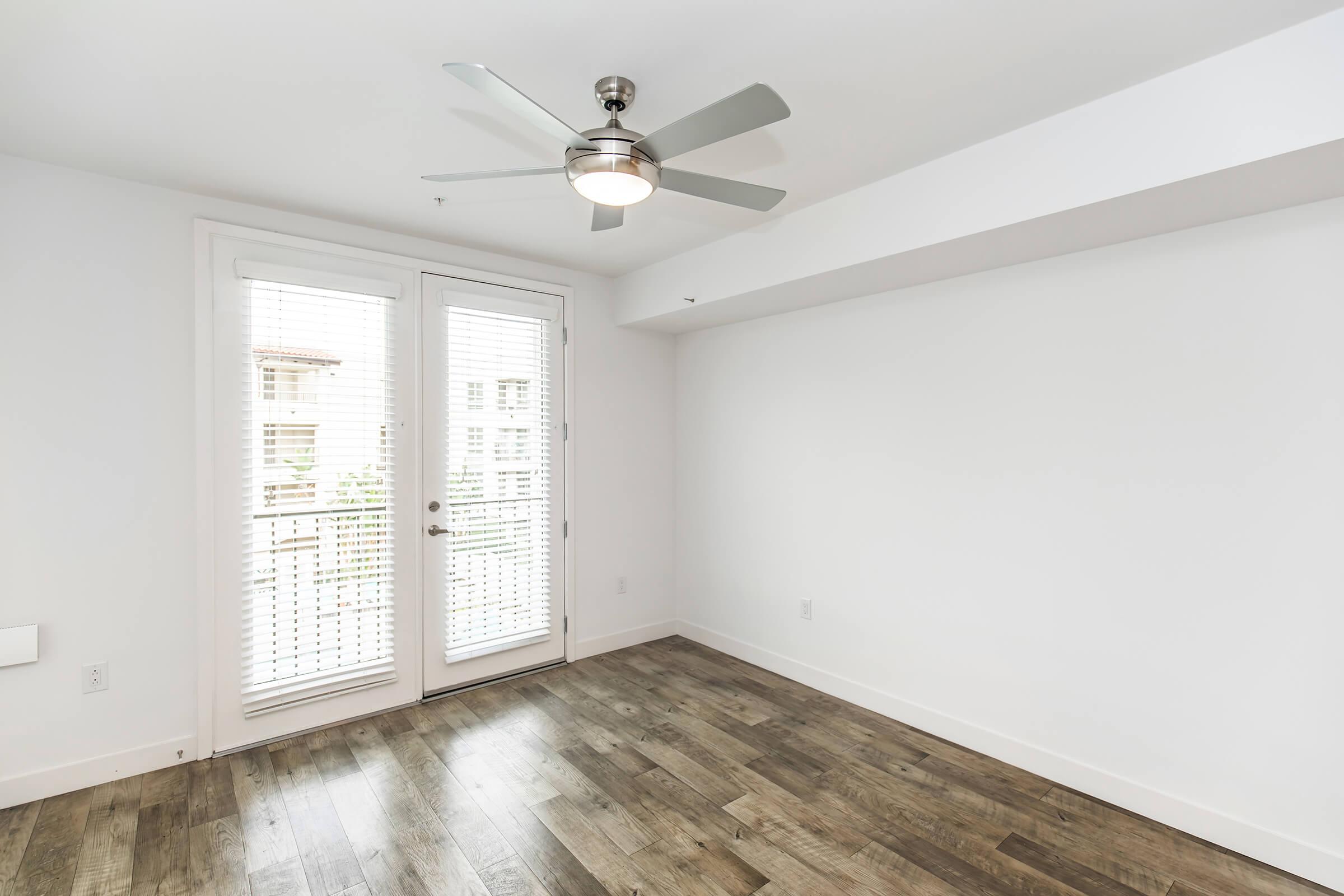 A bright, empty room featuring a ceiling fan and large windows with blinds. The floor is made of wood, and there is a balcony door leading outside. The walls are painted white, creating an open and airy atmosphere.