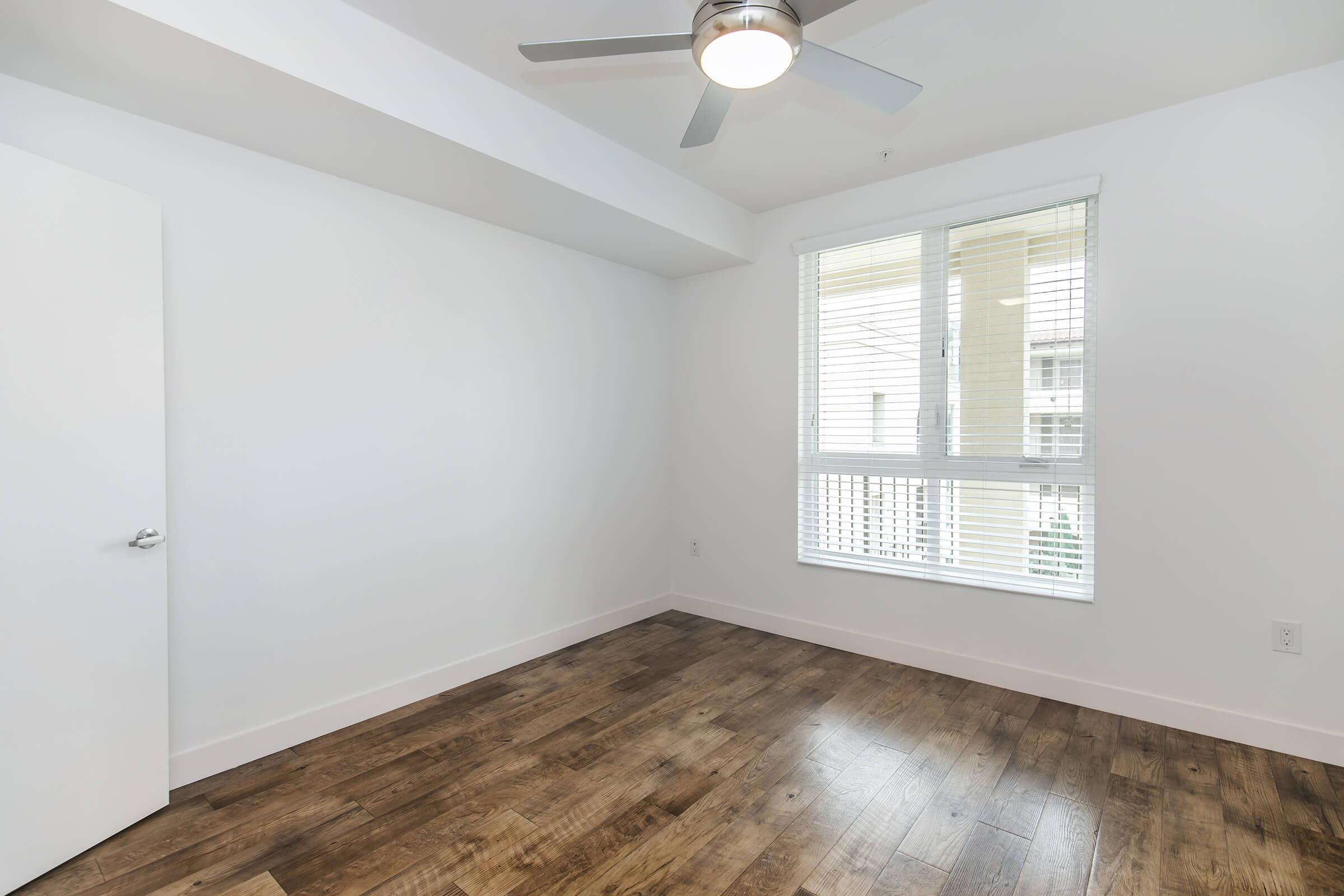 A minimalist room with white walls and a ceiling fan. The floor is made of wood, and there is a window with blinds letting in natural light. A closed white door is visible on the left side, and the overall space appears empty and bright, suitable for various uses.