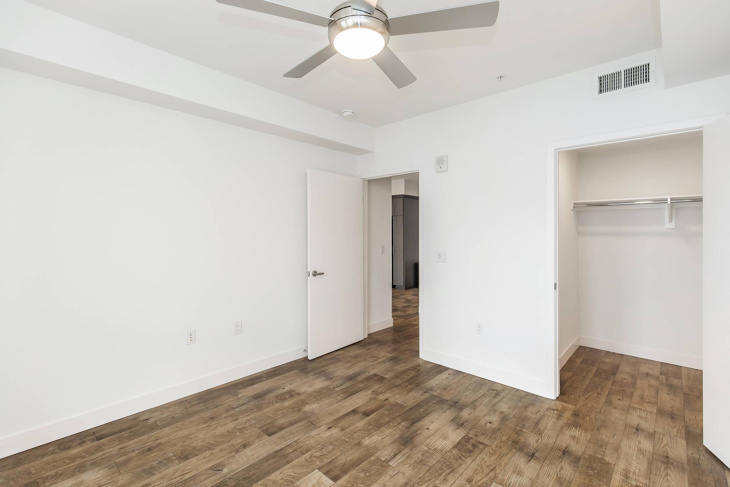 A spacious, modern room with white walls and a ceiling fan. The floor is made of wood, adding warmth to the space. A closet is visible on the right with a door leading to another area. Natural light can flow in through the open doorway, creating a bright and airy atmosphere.