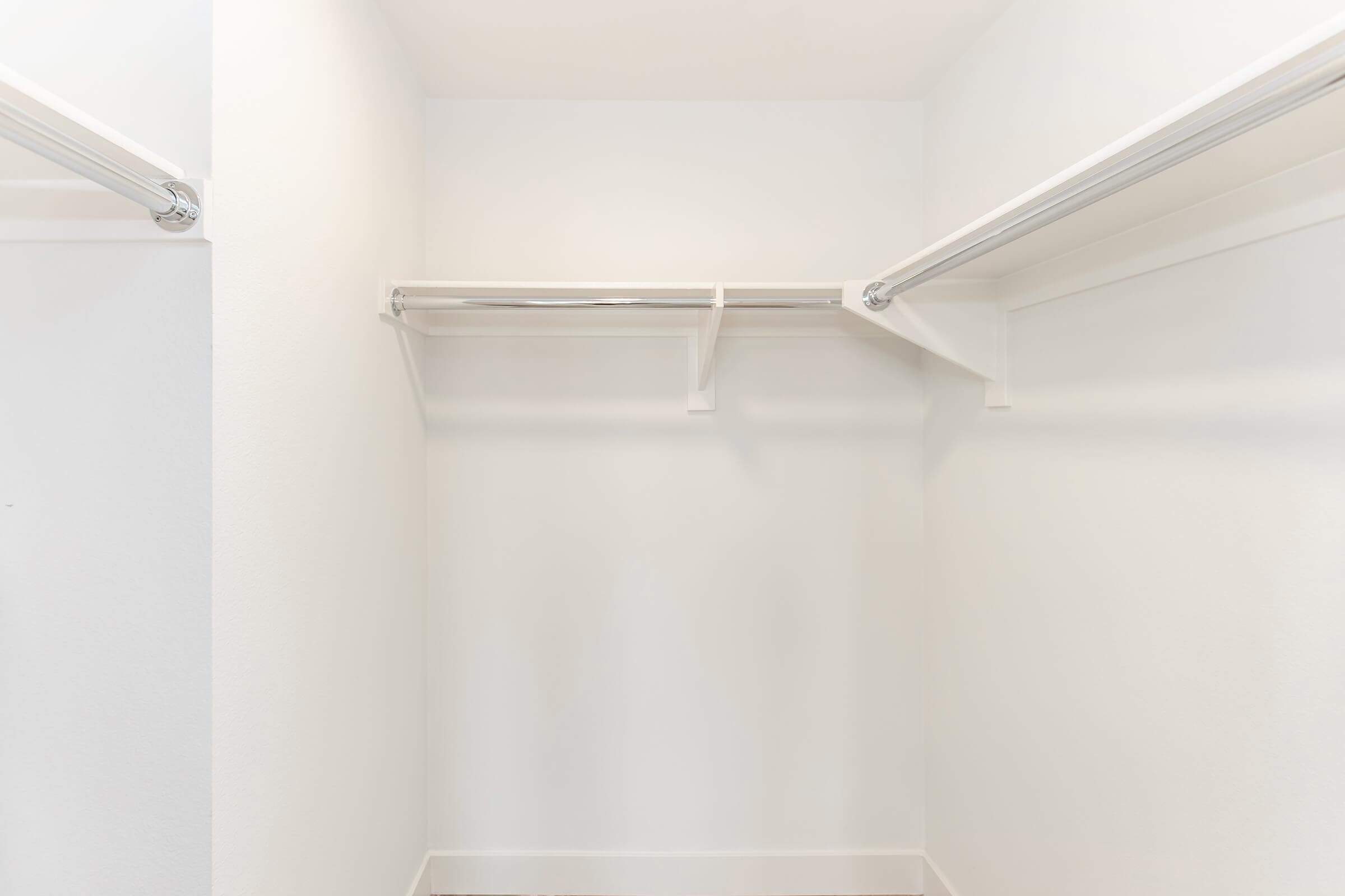 A clean, empty closet with white walls and a wooden floor. Two horizontal clothing rods are mounted on the walls, and a single shelf is positioned above them, also in white. The overall appearance is bright and minimalistic, suggesting a spacious storage area.