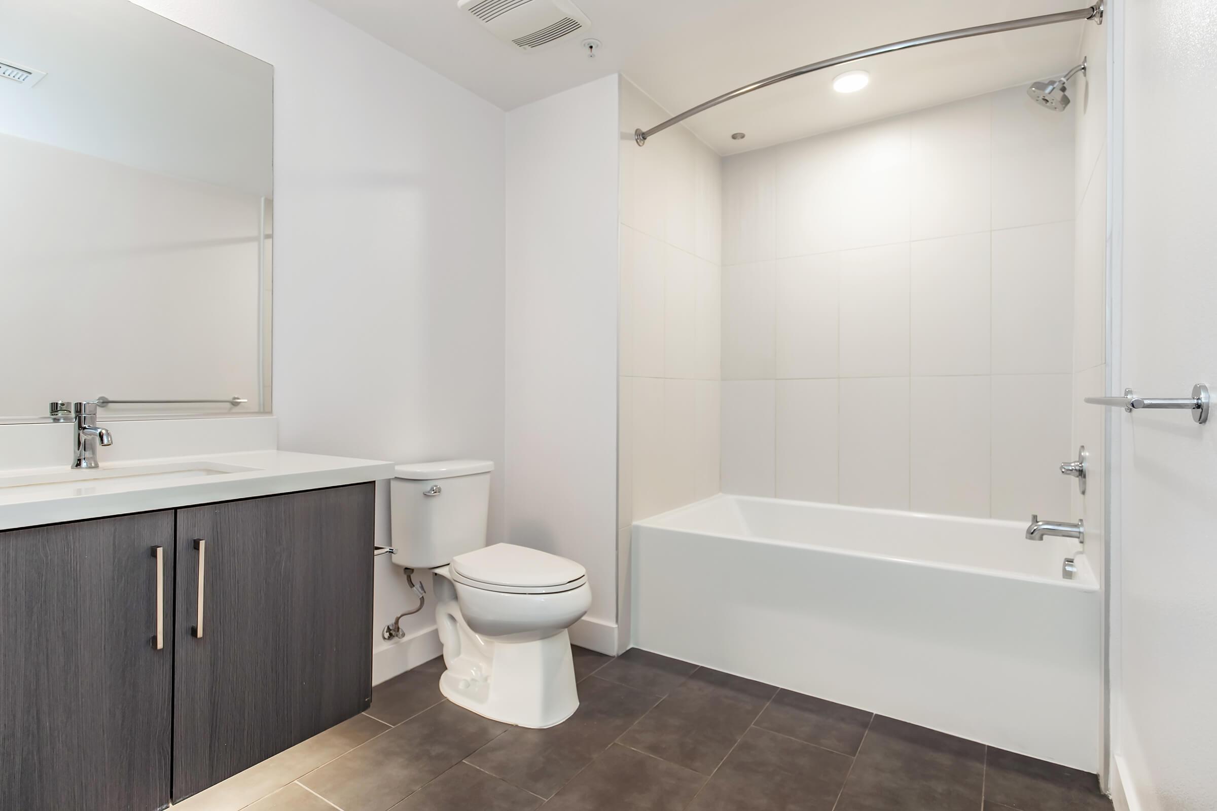 Modern bathroom featuring a white bathtub, shower with a glass curtain, a wall-mounted mirror, a sleek sink with a dark wood cabinet, and a toilet. The walls are finished in white tiles, and the floor is tiled in dark gray, creating a clean and contemporary look.