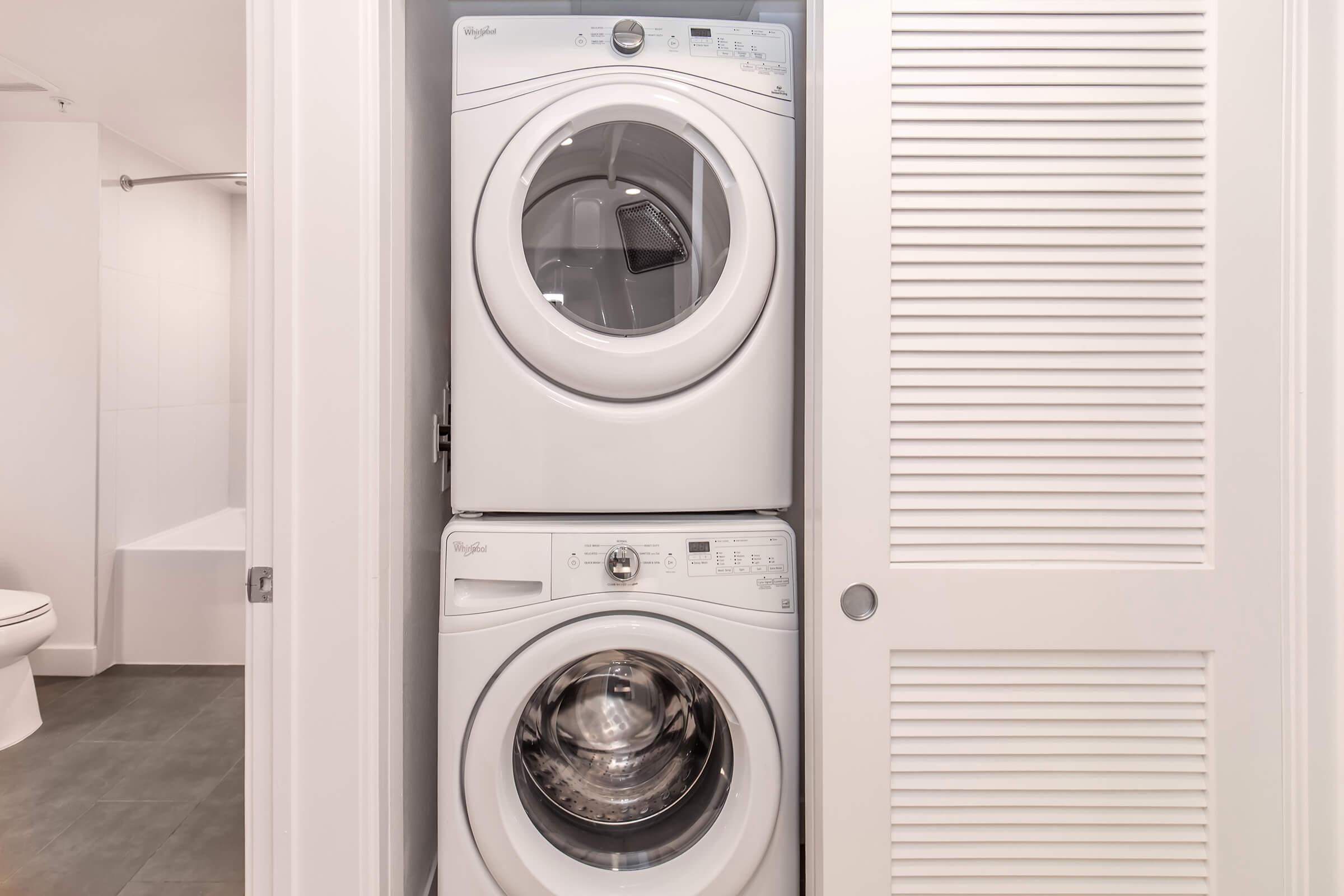 A stacked washer and dryer in a closet, surrounded by white cabinetry. The appliances are modern and white, with the dryer on top and the washer below. In the background, a light-colored bathroom can be seen, featuring a toilet and a partially visible bathtub.