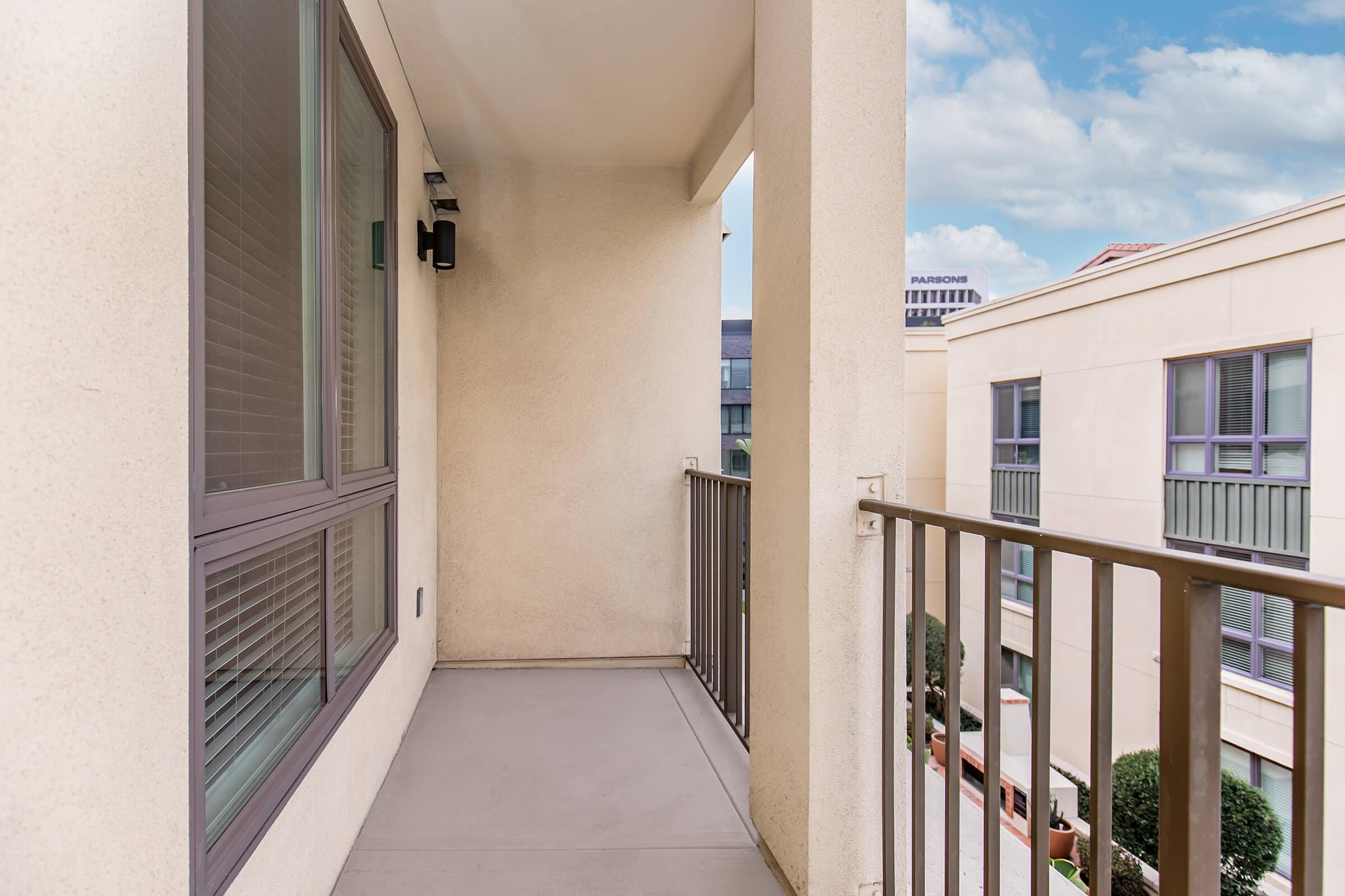 A modern balcony with light-colored walls, featuring a safety railing. The floor is gray, and there are windows with blinds on one side. In the background, part of a building is visible, yielding glimpses of neighboring structures and a clear blue sky.