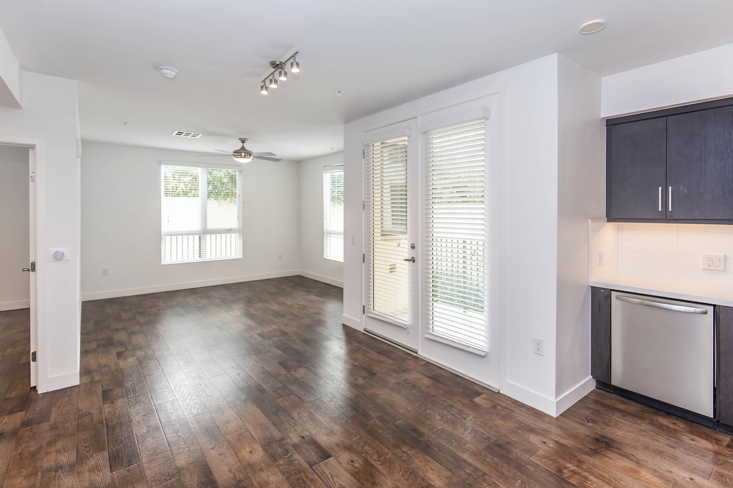 A spacious, modern apartment interior featuring a living area with hardwood floors, large windows, and a ceiling fan. The kitchen is partially visible, showcasing dark cabinetry and stainless steel appliances. The open layout offers plenty of natural light and a welcoming atmosphere.