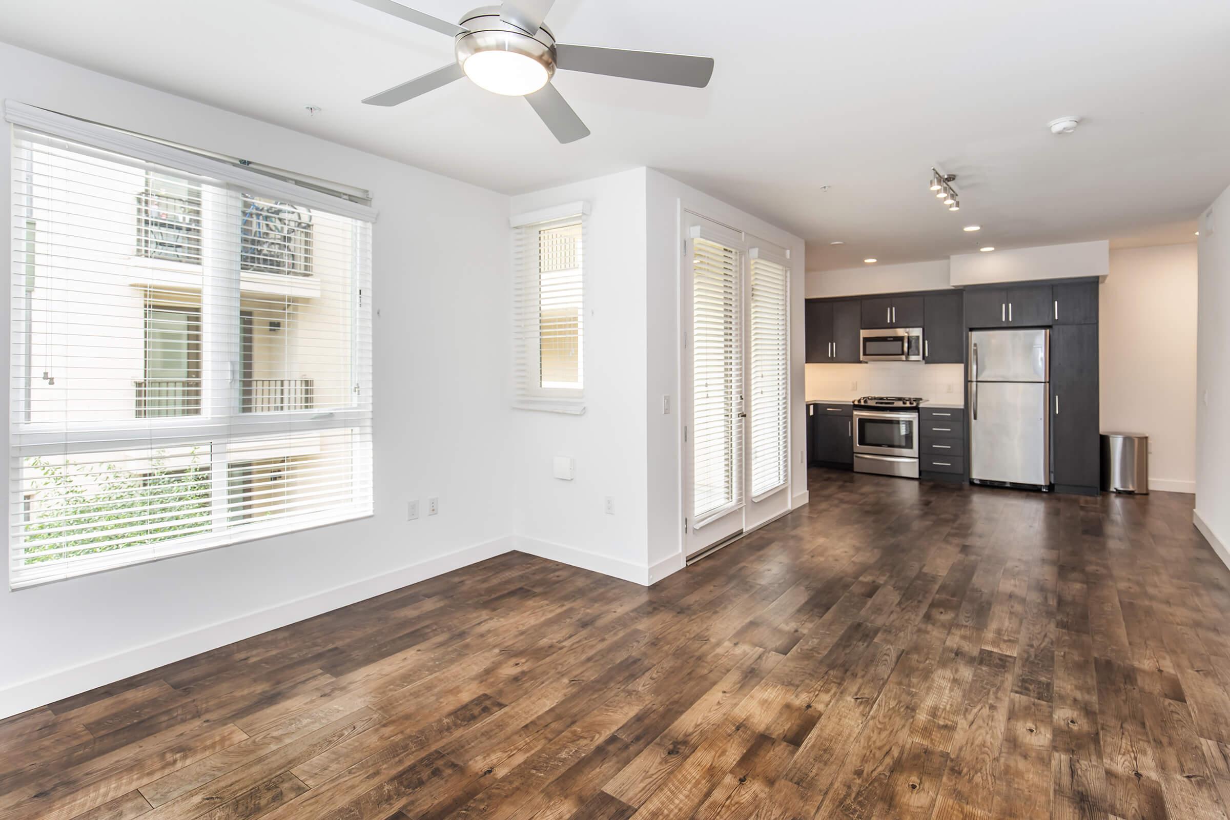 A bright and modern apartment interior featuring an open layout. The living area has large windows with blinds, a ceiling fan, and wooden floors. The kitchen is equipped with stainless steel appliances and dark cabinetry. Natural light fills the space, creating an inviting atmosphere.
