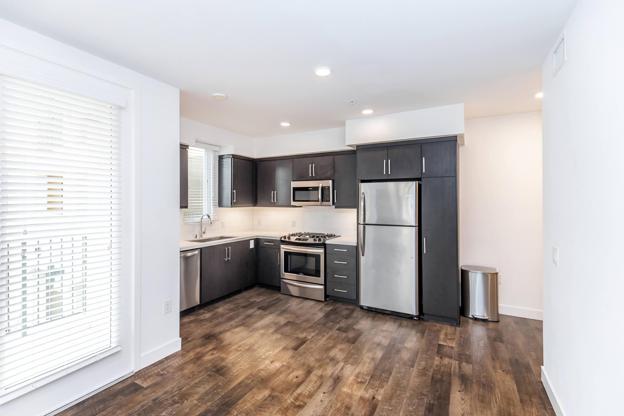 Modern kitchen featuring dark cabinetry, stainless steel appliances, and a gas stove. The space includes white quartz countertops, a window with blinds, and light wood flooring. A stainless steel trash can is visible in the corner, contributing to a clean and contemporary look.