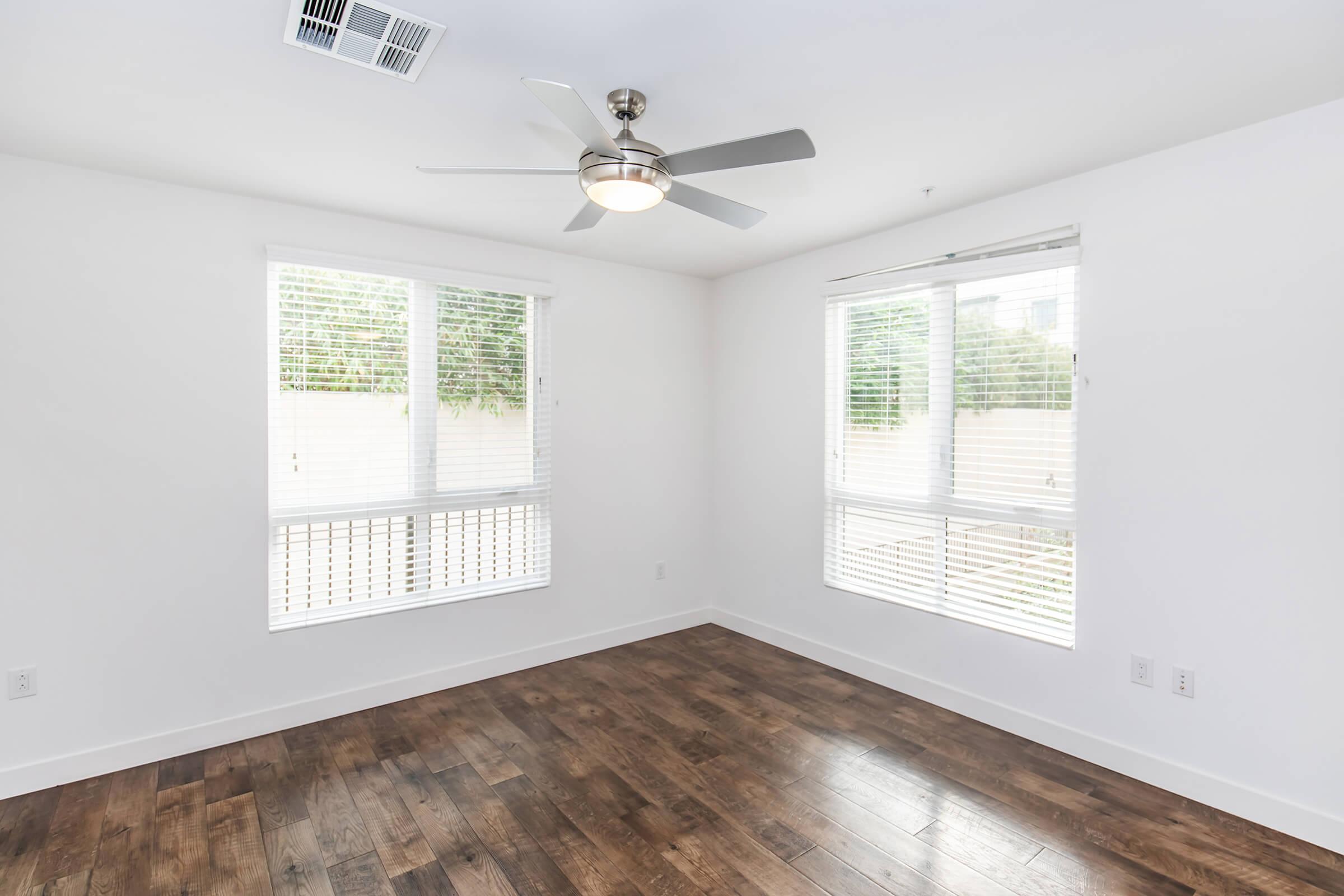 Bright, modern room featuring large windows with white blinds, a ceiling fan, and hardwood flooring. The walls are painted white, creating a clean and spacious atmosphere.
