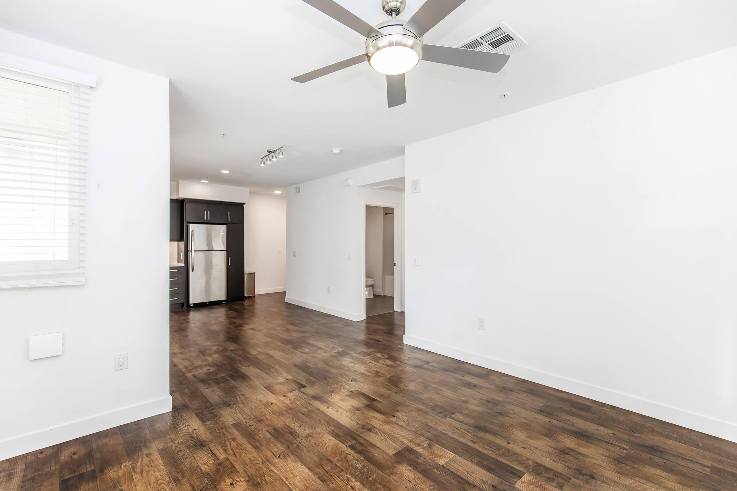 A modern, open-concept living space featuring light-colored walls, hardwood flooring, and a ceiling fan. The kitchen area includes stainless steel appliances and dark cabinetry. Natural light enters through a window with blinds, creating a bright and airy atmosphere.