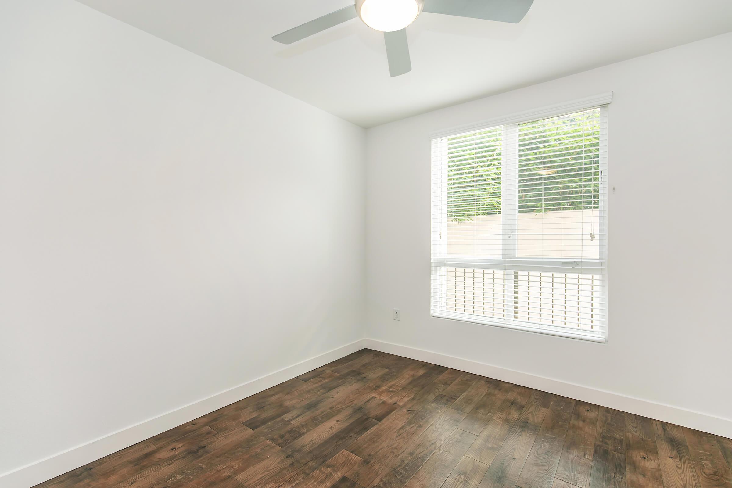 Bright and empty room with white walls and a wooden floor. A ceiling fan is visible, and a large window with closed blinds allows natural light to enter the space. The overall atmosphere is clean and minimalist, suitable for various uses.