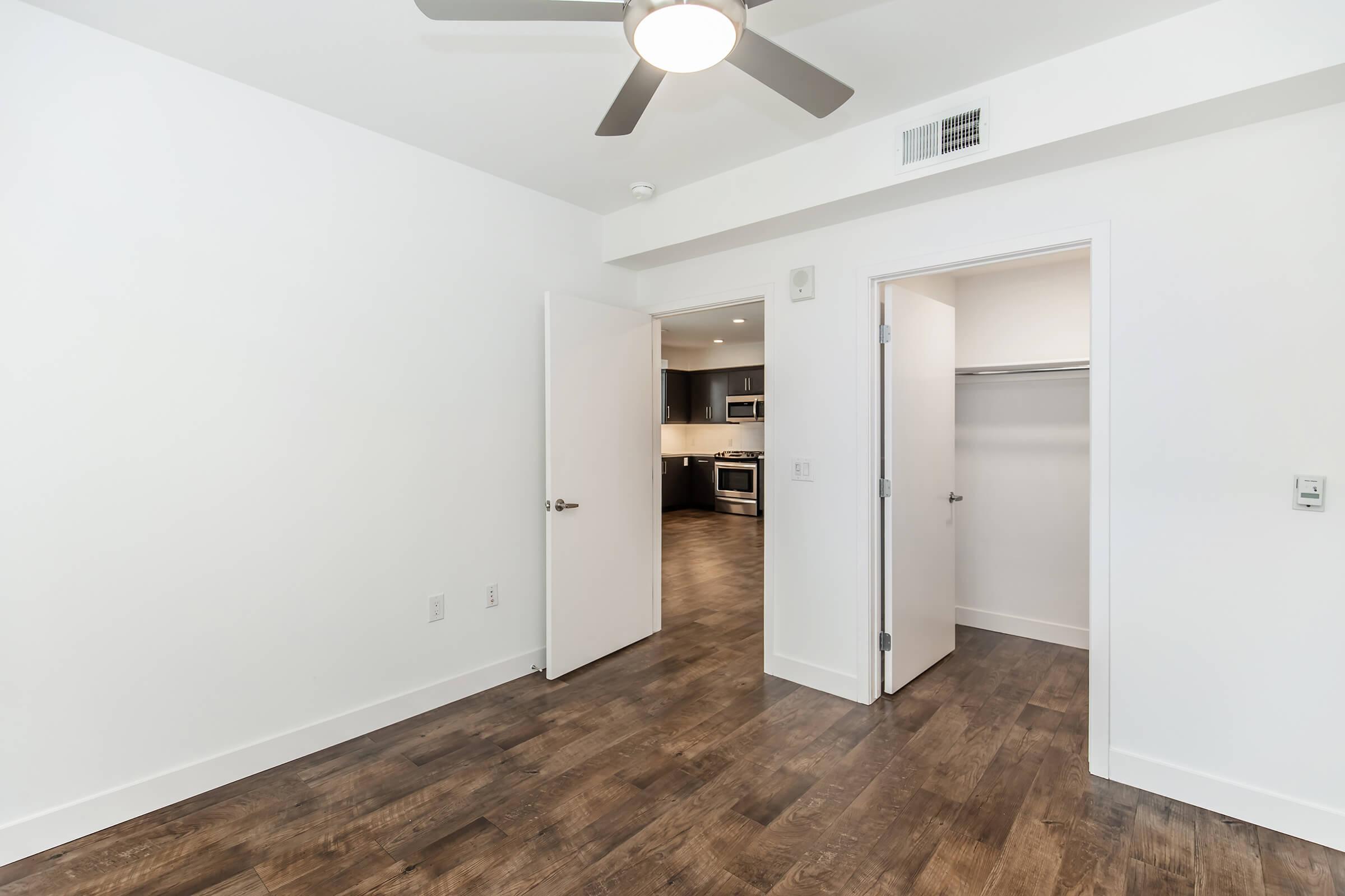 An empty room with white walls and a ceiling fan, featuring open doors leading to a kitchen area and a closet. The floor is made of dark wood. Natural light highlights the spacious layout, creating a clean and modern ambiance.