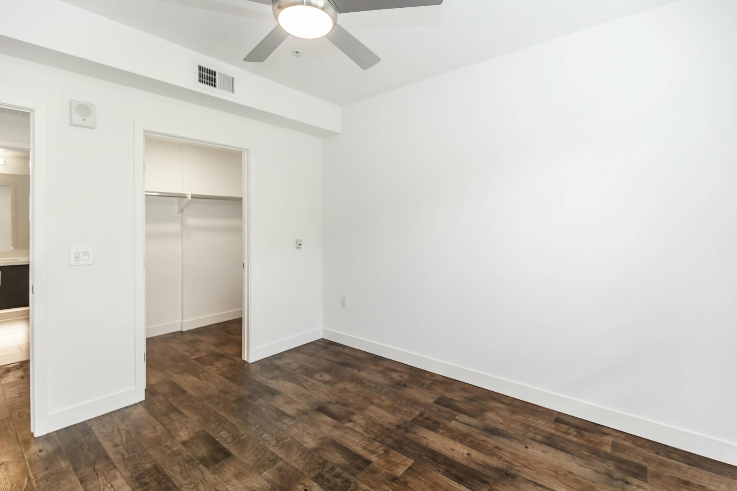 A spacious, empty room with light-colored walls and a hardwood floor. On one side, there is a doorway leading to a closet with an open hanging space. A ceiling fan is mounted in the center, and soft natural light illuminates the space, giving it a bright and airy feel.