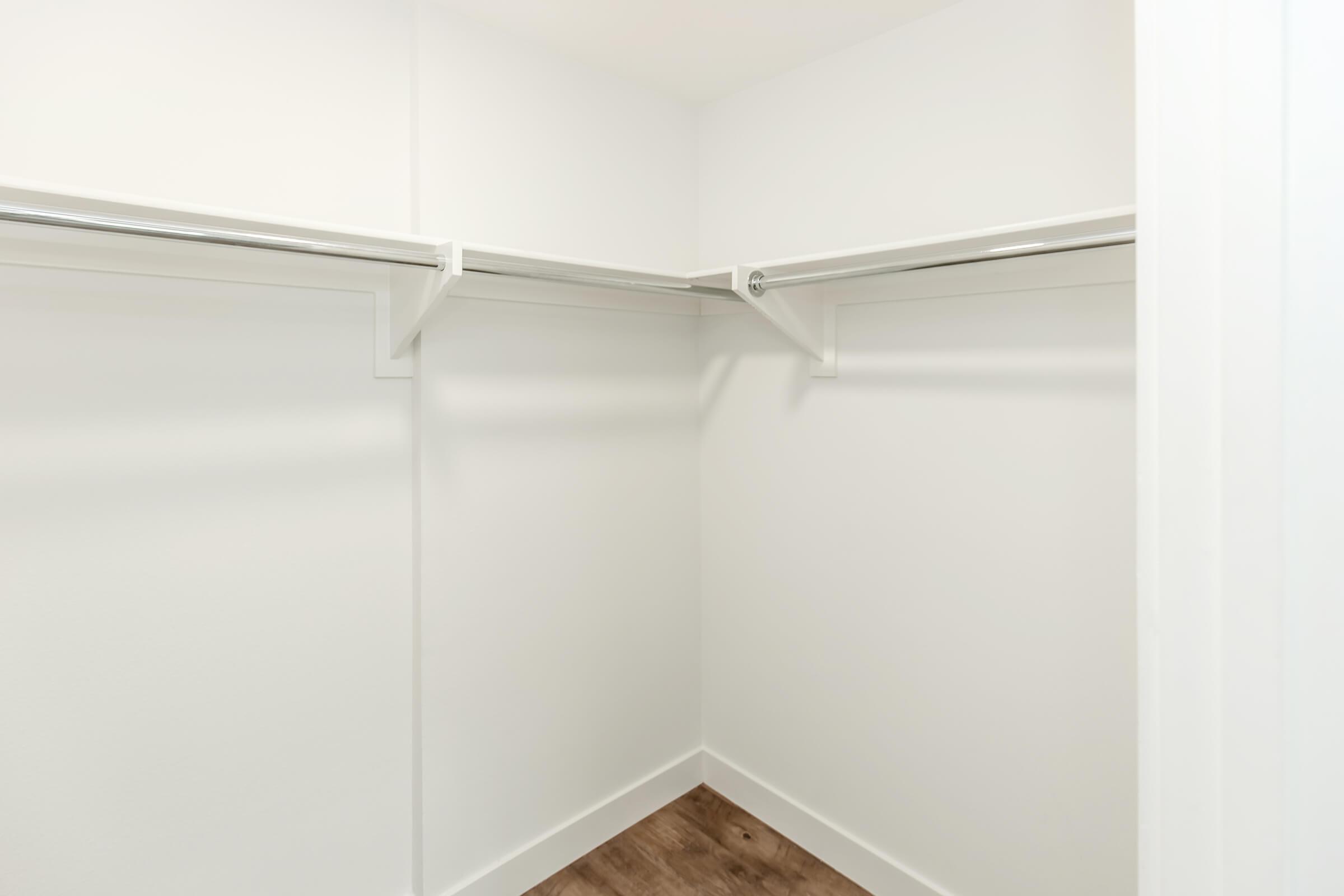 Empty closet with white walls and shelves, featuring two horizontal clothes rods for hanging items. The floor is a light wood finish, creating a bright and minimalistic storage space.