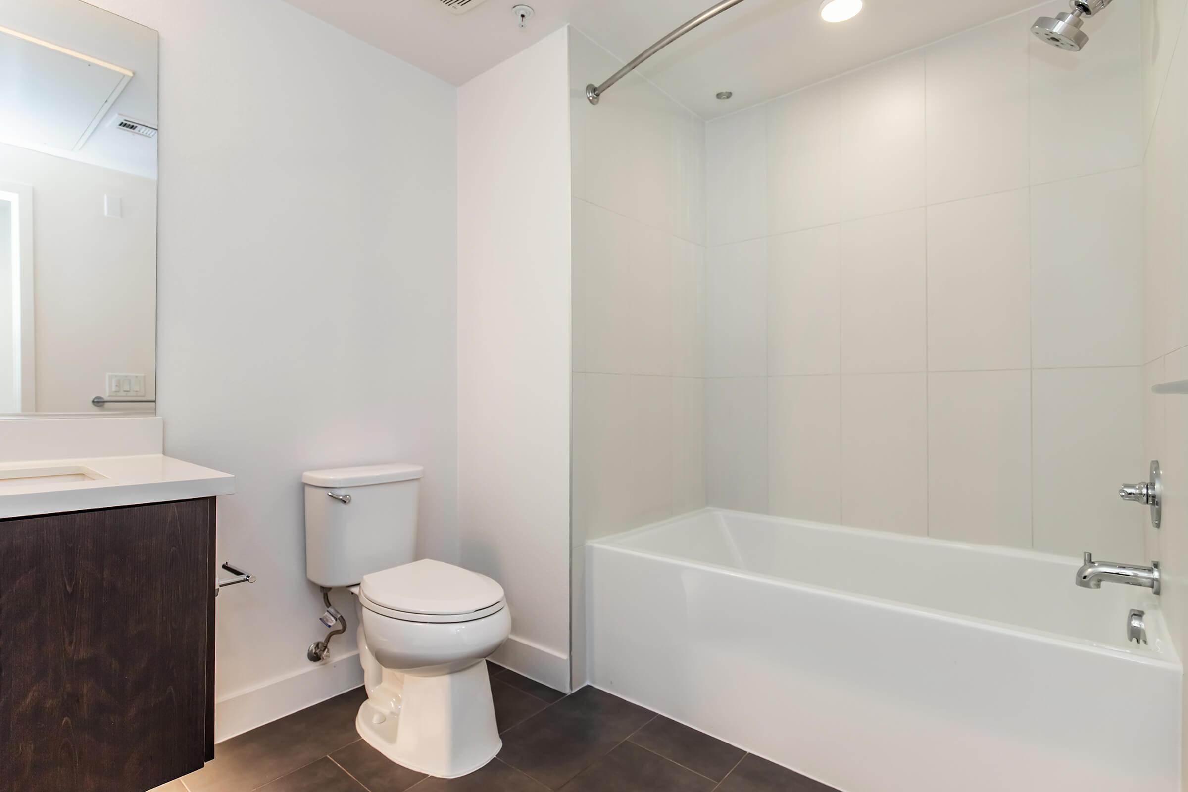 A modern bathroom featuring a white bathtub with a shower, a wall-mounted mirror above a sink, and a white toilet. The walls are tiled in light colors, and the floor has dark tiles, creating a contemporary atmosphere. Bright lighting enhances the clean and spacious look of the room.