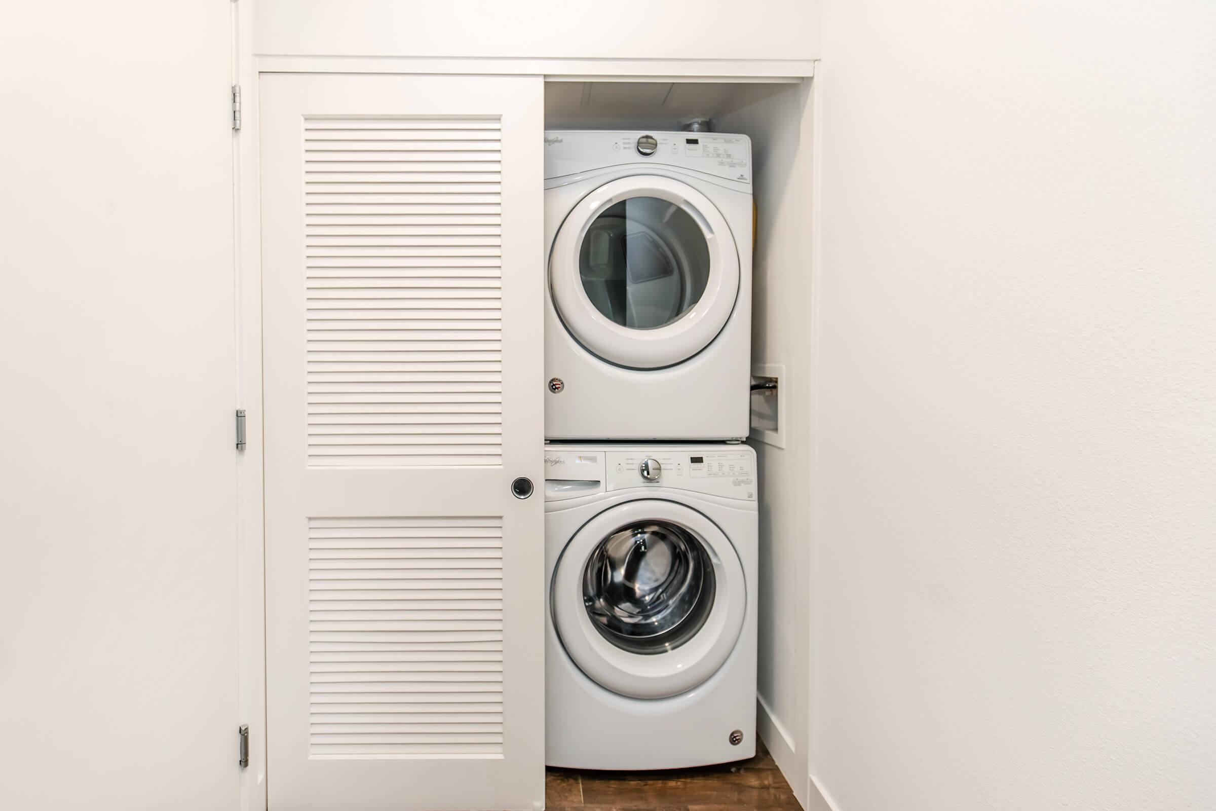 A stacked washer and dryer unit situated in a small closet with white shutter doors, against a plain white wall. The appliances are modern and both feature large circular doors. The floor is made of wood, adding warmth to the otherwise minimalistic space.