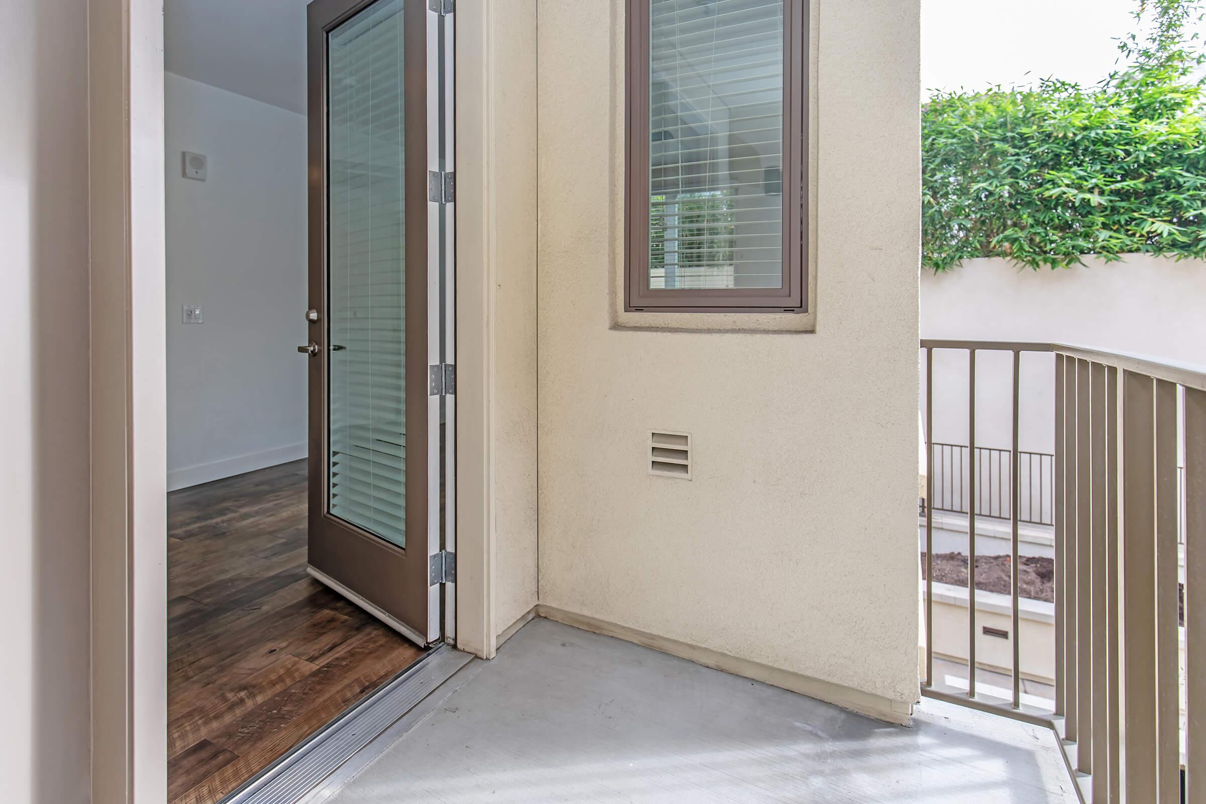 A doorway opening to a small balcony or outdoor area with a railing. The interior walls are pale, and there are large windows allowing natural light. The floor is wooden, and there are green plants visible outside, creating a pleasant view.