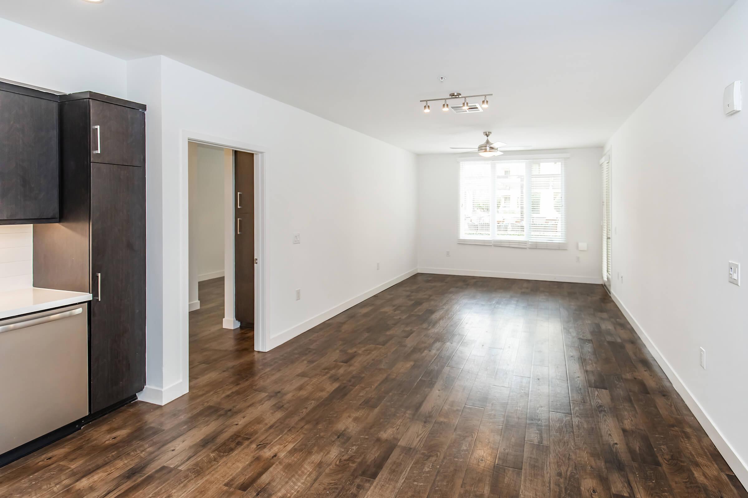 A modern, empty living space with hardwood floors, featuring a large window that lets in natural light. The kitchen area is visible on the left with dark cabinetry and stainless steel appliances. There is a doorway leading to another room, adding to the open, spacious feel of the area.