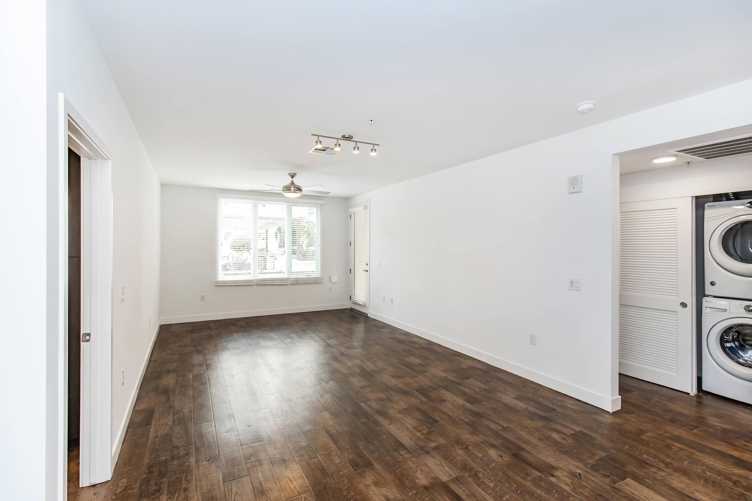 A bright and spacious living area featuring large windows, wood flooring, and a ceiling light fixture. On the right side, there is a stackable washer and dryer unit in a closet. The walls are painted white, creating a clean and modern aesthetic.