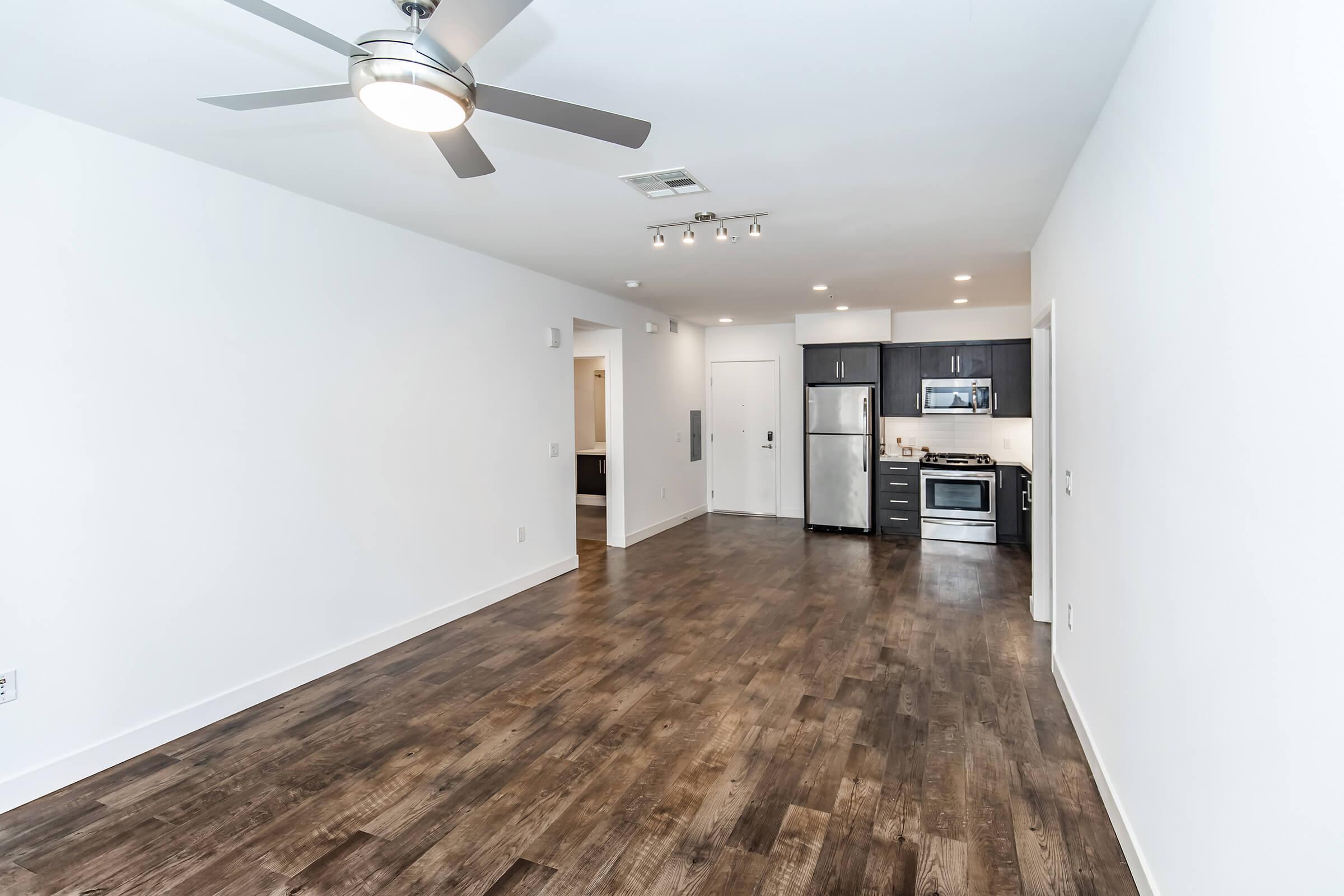 A spacious, modern apartment interior featuring a living area with hardwood floors, a ceiling fan, and an open kitchen. The kitchen includes stainless steel appliances, dark cabinetry, and a countertop. The walls are painted white, creating a bright and airy atmosphere.