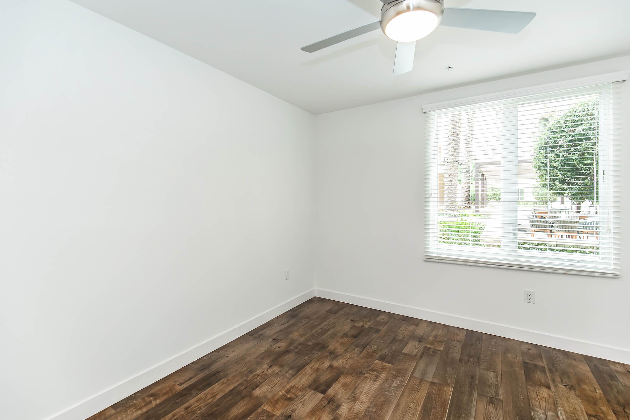 A bright, empty room with light-colored walls and a wooden floor. A ceiling fan is mounted above, and a window with white blinds allows natural light to fill the space, showcasing greenery outside. The room is unadorned, creating a minimalist and open atmosphere.