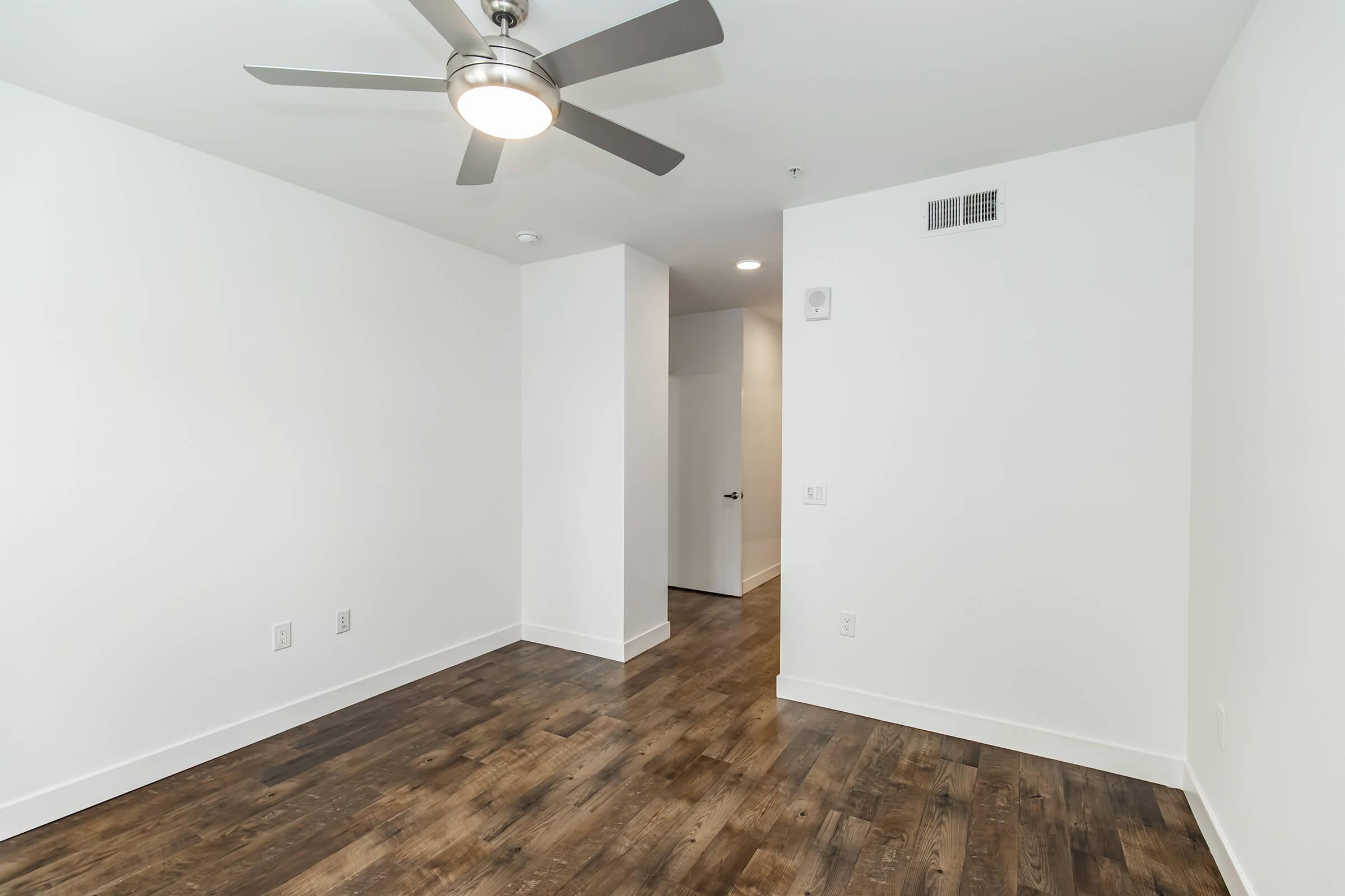 A spacious, empty room featuring a ceiling fan, white walls, and dark wooden flooring. The room has a doorway leading to another area and modern lighting. The overall aesthetic is clean and minimalistic, ideal for personal decoration.