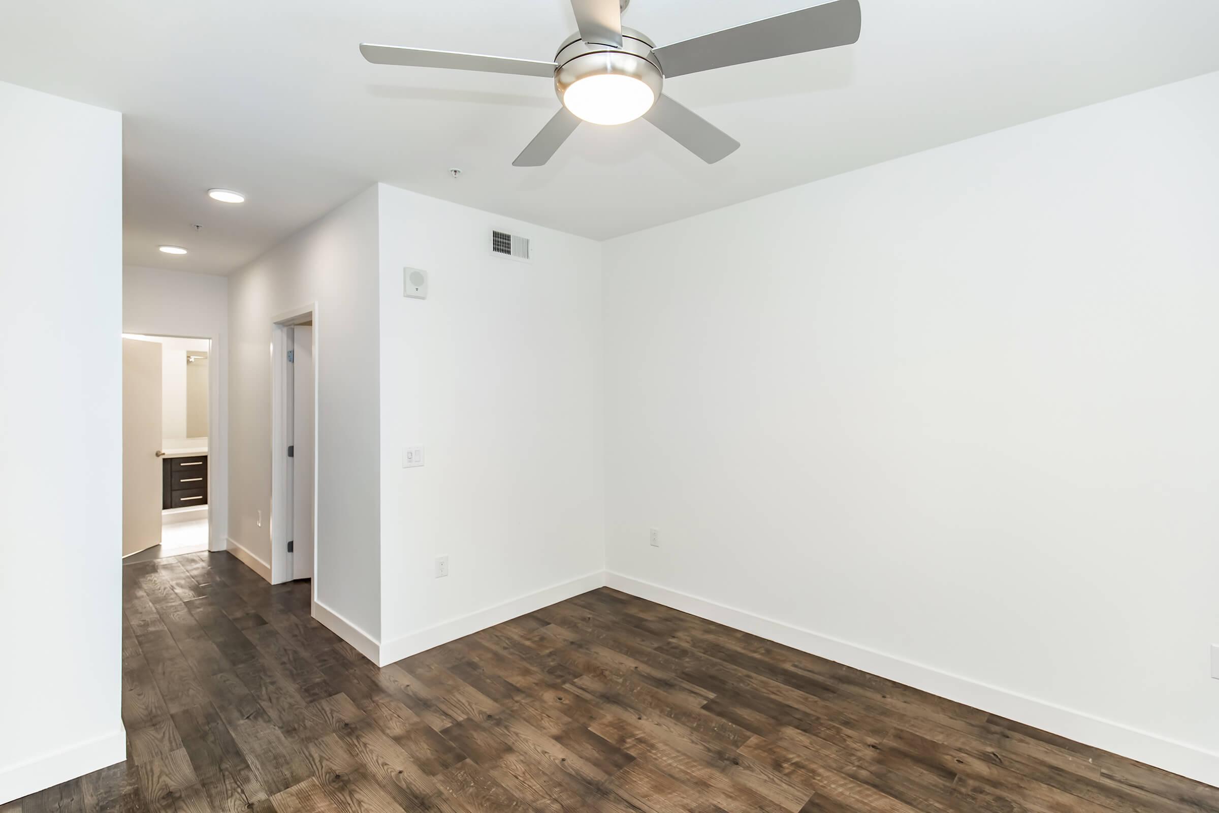 A bright, empty room featuring a ceiling fan and modern light fixtures. The floor is made of wooden planks, and there are white walls. A doorway leads to a bathroom visible in the background, with a sleek, contemporary design.