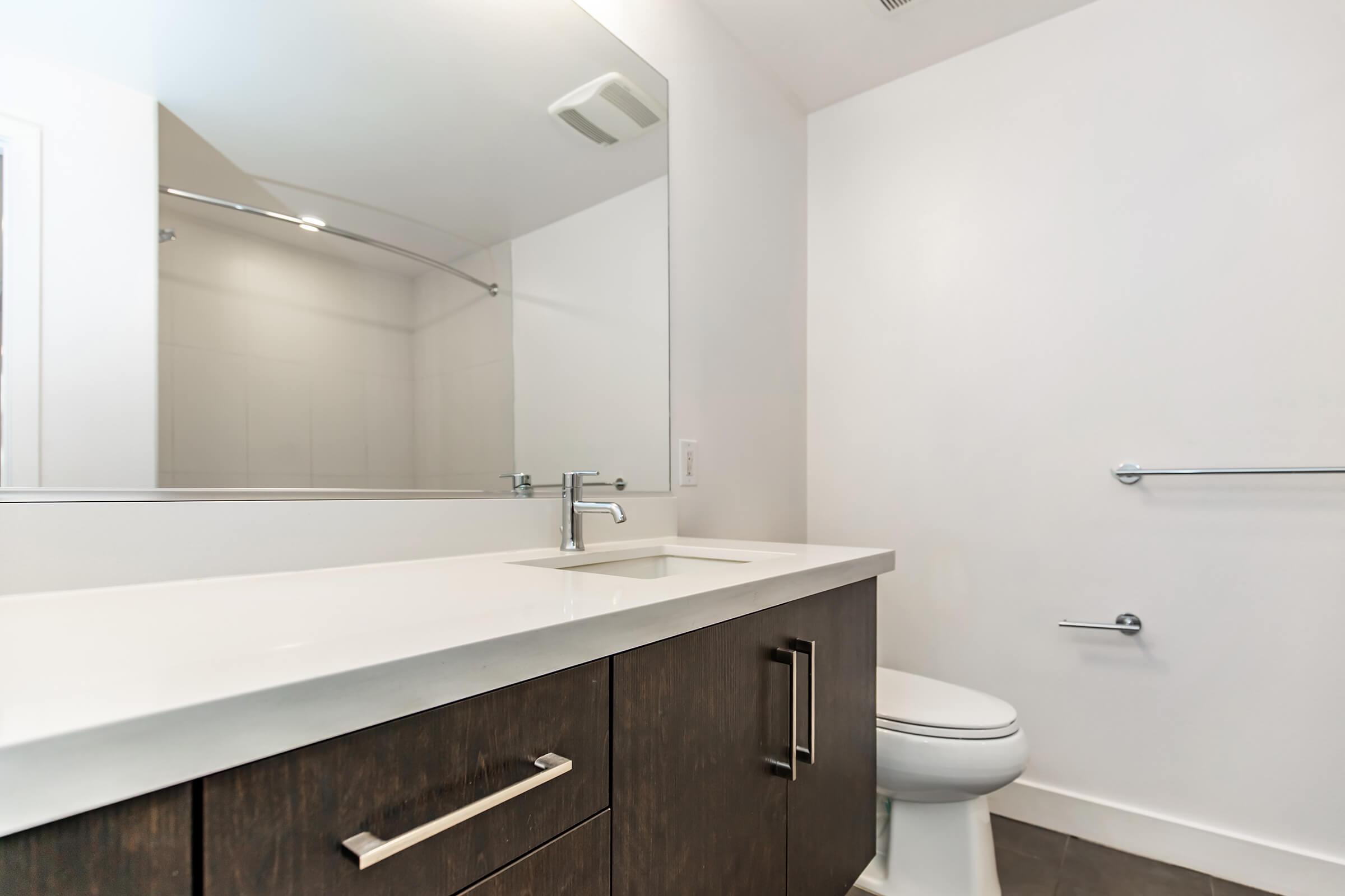 A modern bathroom featuring a clean, minimalist design. It includes a white sink with a sleek faucet, a large mirror above the sink, and a toilet. The cabinetry is dark wood, contrasting with the light-colored walls and floor. A glass shower enclosure is visible in the background.
