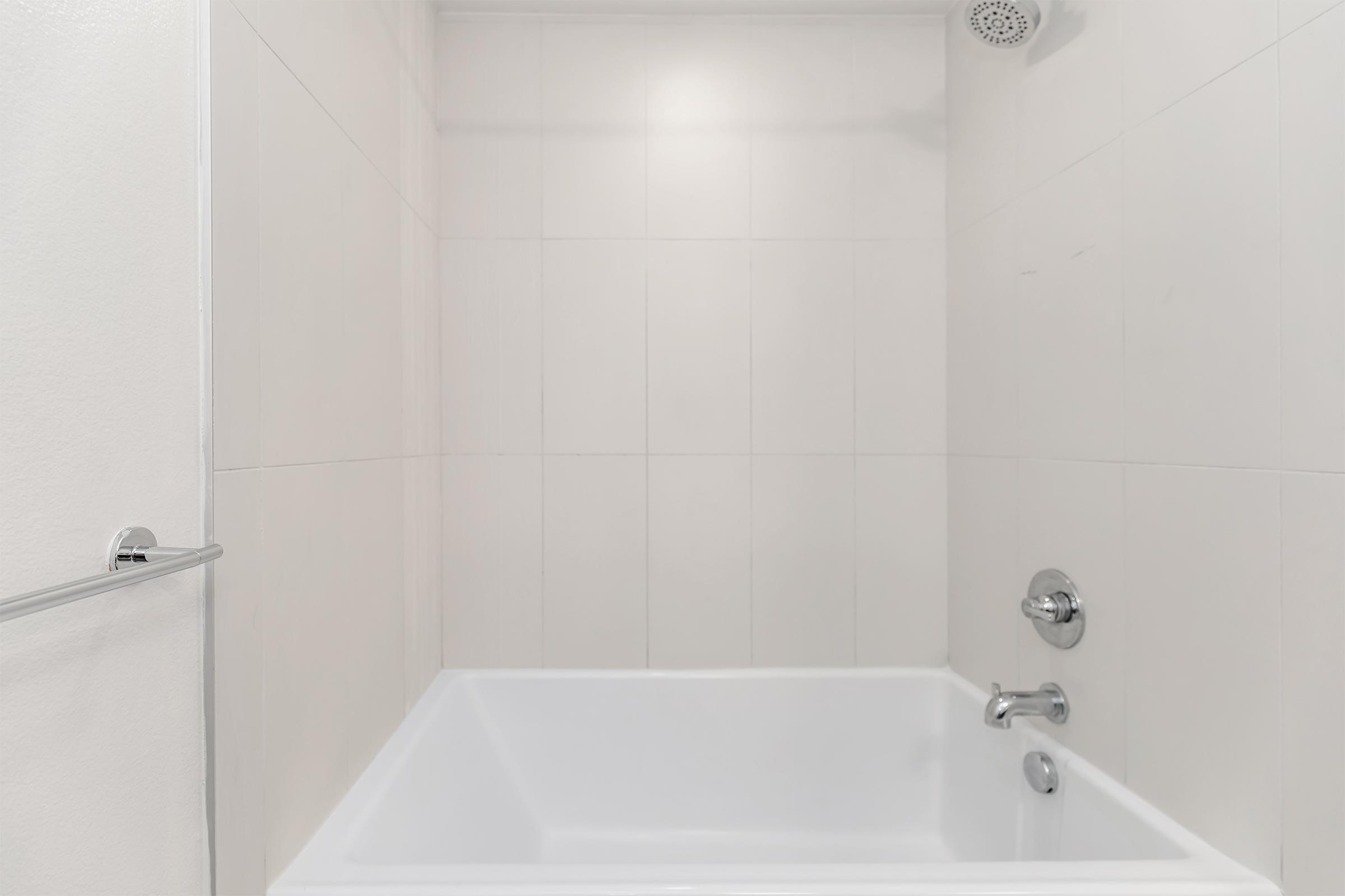 A clean, modern bathroom featuring a shower area with light-colored tile walls, a white bathtub, and a silver faucet. The design emphasizes simplicity and functionality, with a towel bar mounted on the wall and a showerhead above.