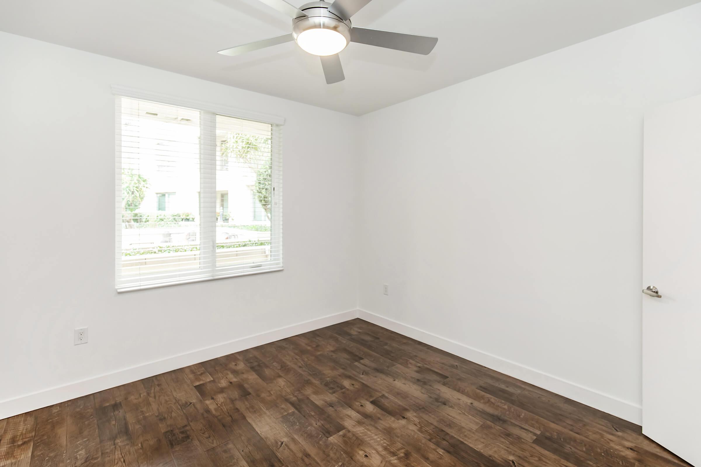 A vacant room featuring a ceiling fan, large window with blinds, and hardwood flooring. The walls are painted white, and there is a simple door on one side. Natural light illuminates the space, enhancing its clean and modern appearance.