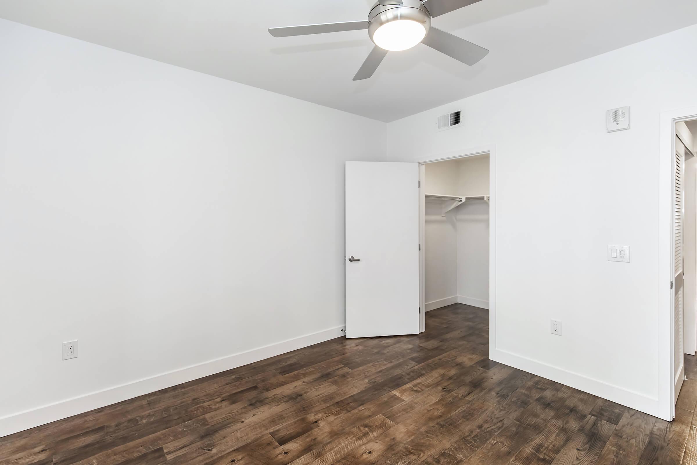 A spacious, modern bedroom with white walls, a ceiling fan, and hardwood flooring. A white door is slightly ajar, leading to a closet with built-in shelving. Natural light fills the room, creating a clean and inviting atmosphere.