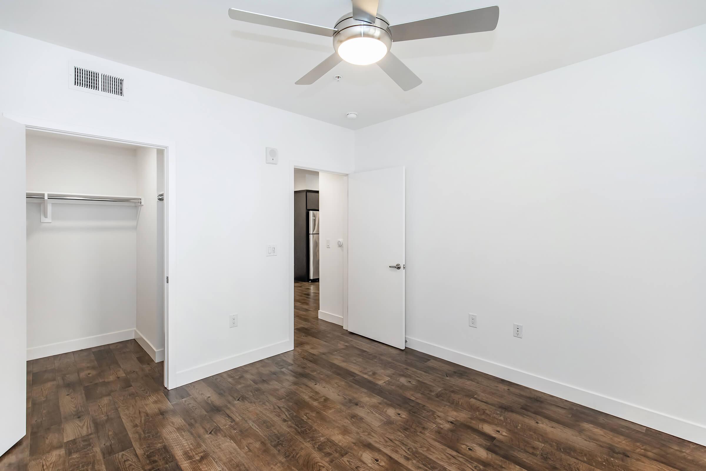 Empty room with white walls, a ceiling fan, and laminate flooring. One wall features a closet without doors, while another has a closed door. A modern and minimalistic design creates a spacious feel.