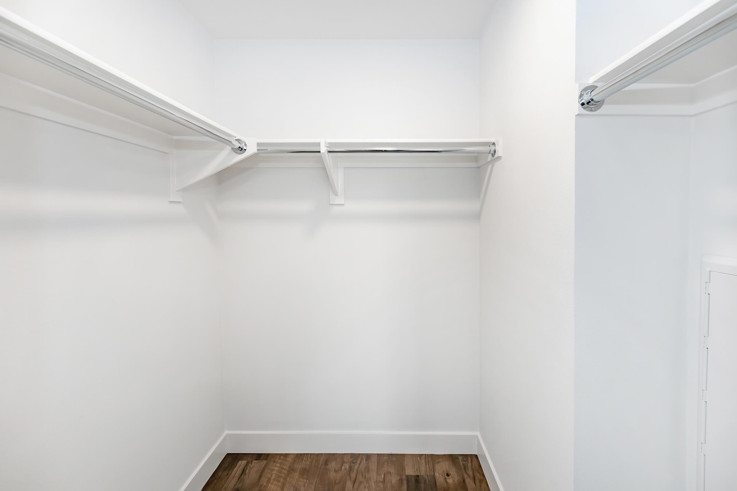 A spacious, empty closet with two shelving rods mounted on the walls, white walls, and a wooden floor. The closet has no visible items, creating a clean and minimalist look, perfect for storage or organization.