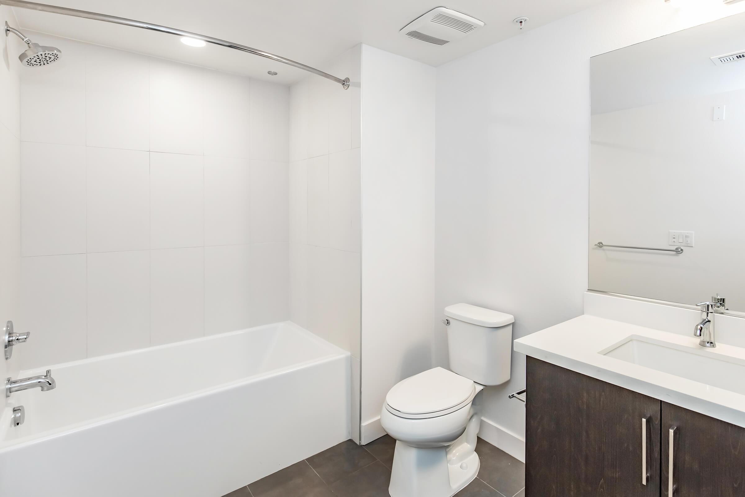 A modern bathroom featuring a white bathtub with a shower curtain rod, a toilet, and a sleek vanity with a mirror. The walls are tiled in light colors, and the flooring is dark. The space appears clean and contemporary.