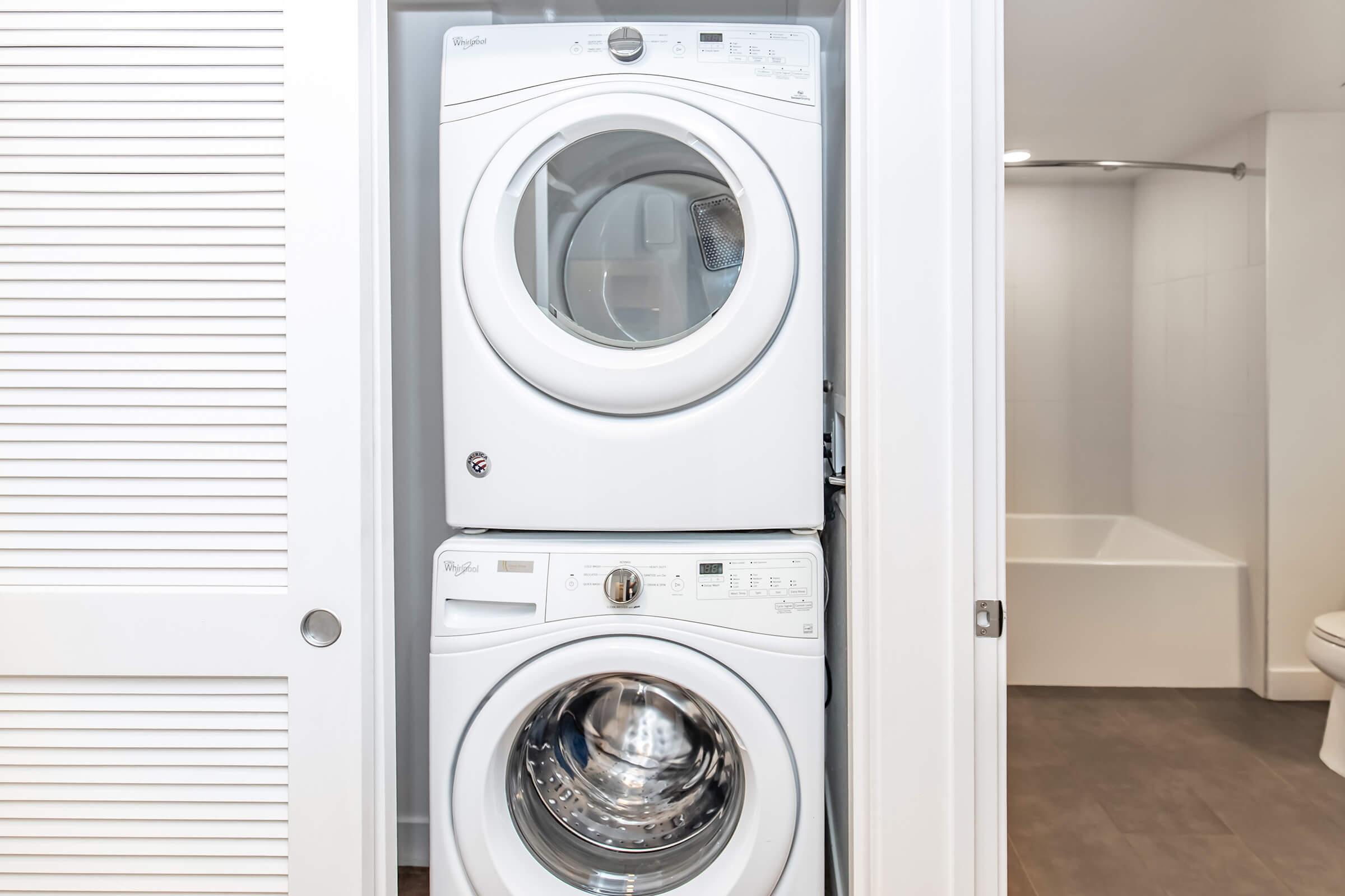 A stacked washer and dryer set in a small laundry area. The machines are white and modern, with the washer on top and the dryer below. The background features a partially open closet door and a bathroom with a bathtub and toilet visible beyond. The overall setting is clean and minimalist.