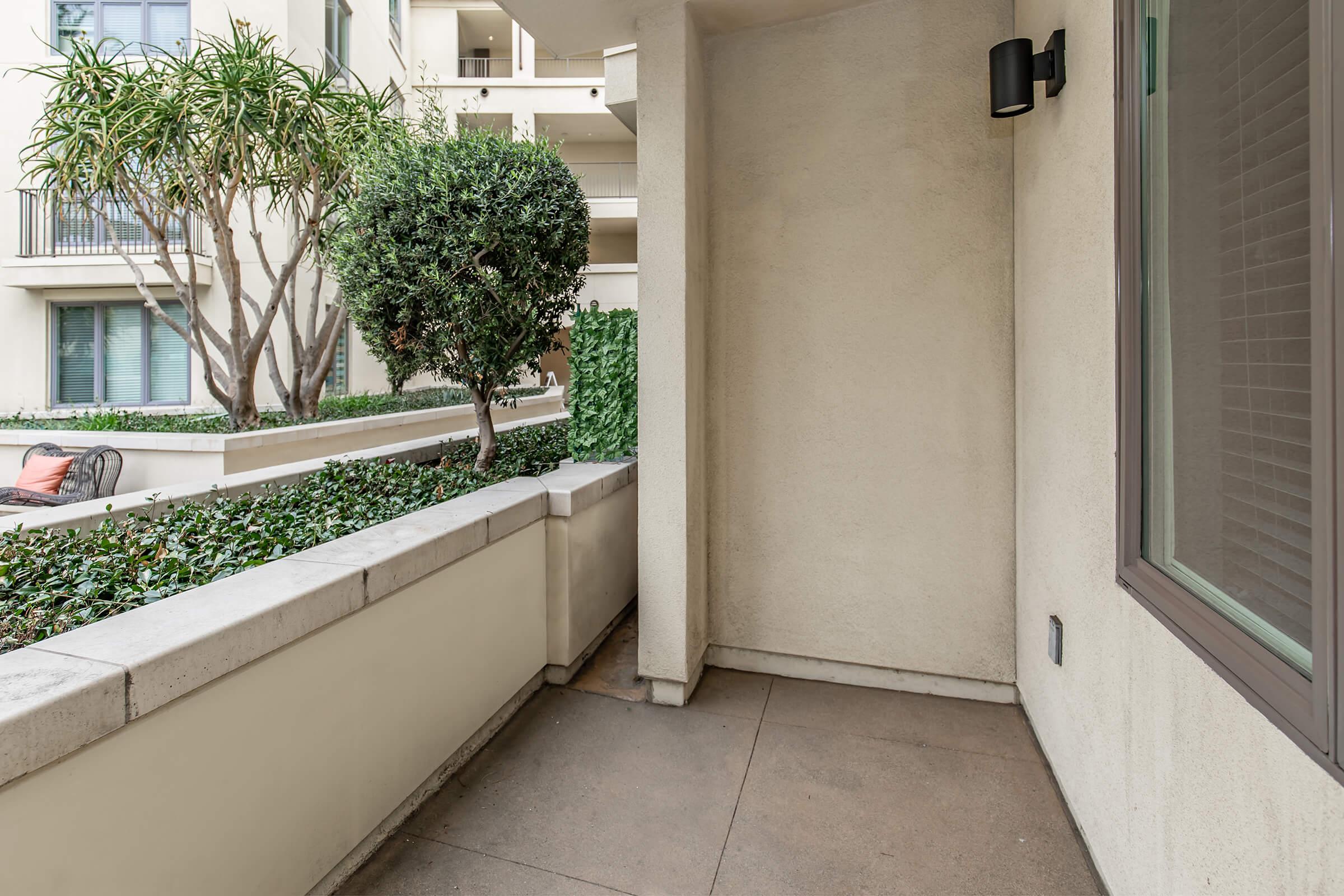 A corner of a balcony or patio featuring potted plants and decorative greenery. The space includes a smooth concrete floor and a wall with a window. There are trees and shrubs visible, surrounded by a modern architectural design of a building.