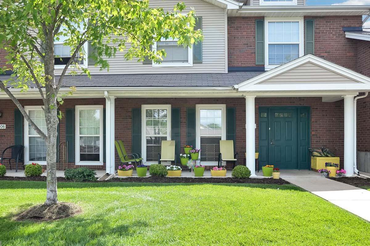 A welcoming front porch featuring green rocking chairs, colorful flower pots, and neatly manicured grass. The brick facade of the house is complemented by white trim and large windows, creating a bright and inviting entrance. A small tree adds greenery to the scene.