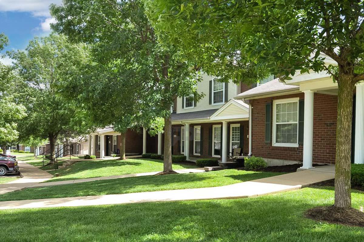 A row of residential buildings with brick and siding exteriors, set among lush green lawns. The scene features trees providing shade and a paved pathway leading through the neighborhood. The sky is bright with a few clouds, creating a pleasant outdoor atmosphere.