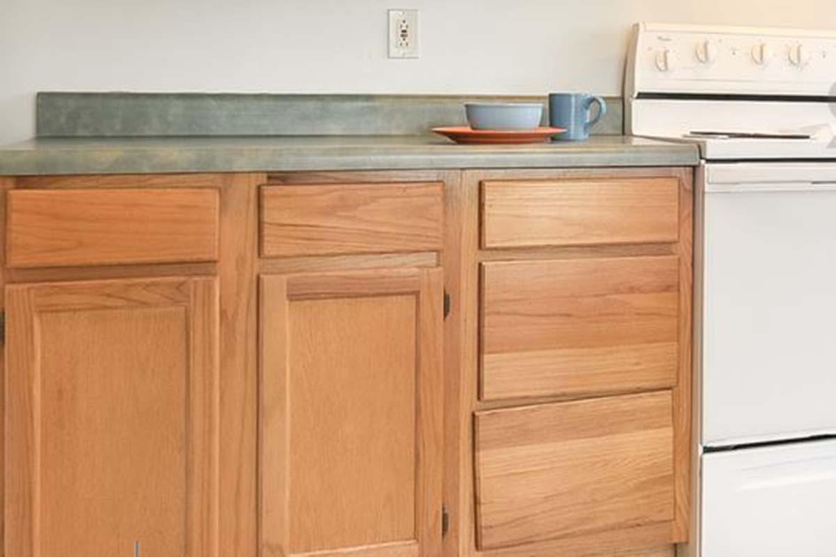 A wooden kitchen cabinet with multiple drawers and doors, featuring a gray countertop. On the countertop, there is a blue bowl, a blue mug, and an orange plate. An oven is located next to the cabinetry. The walls are painted a neutral color.