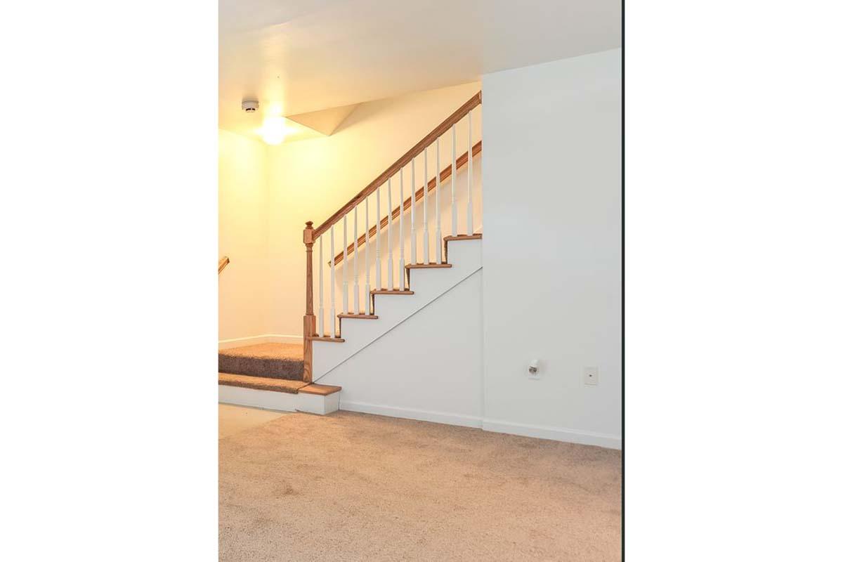 Interior of a home featuring a carpeted area with a staircase in the background. The staircase has wooden railings and steps leading to a higher level. The walls are painted white, and there is soft lighting illuminating the space.