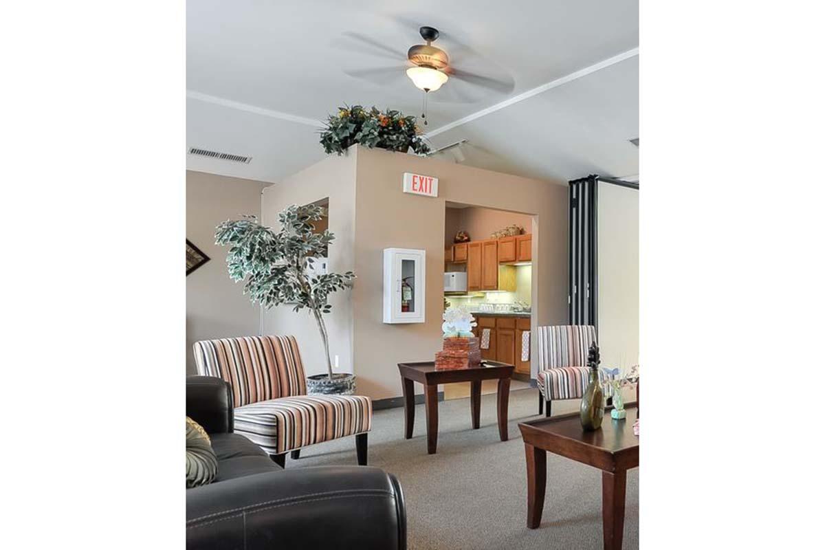 A cozy living space featuring two striped chairs and a wooden table, with a decorative plant nearby. A ceiling fan is overhead, while an exit sign and a partial view of a kitchen area can be seen in the background. The walls are a soft beige color, contributing to a warm atmosphere.