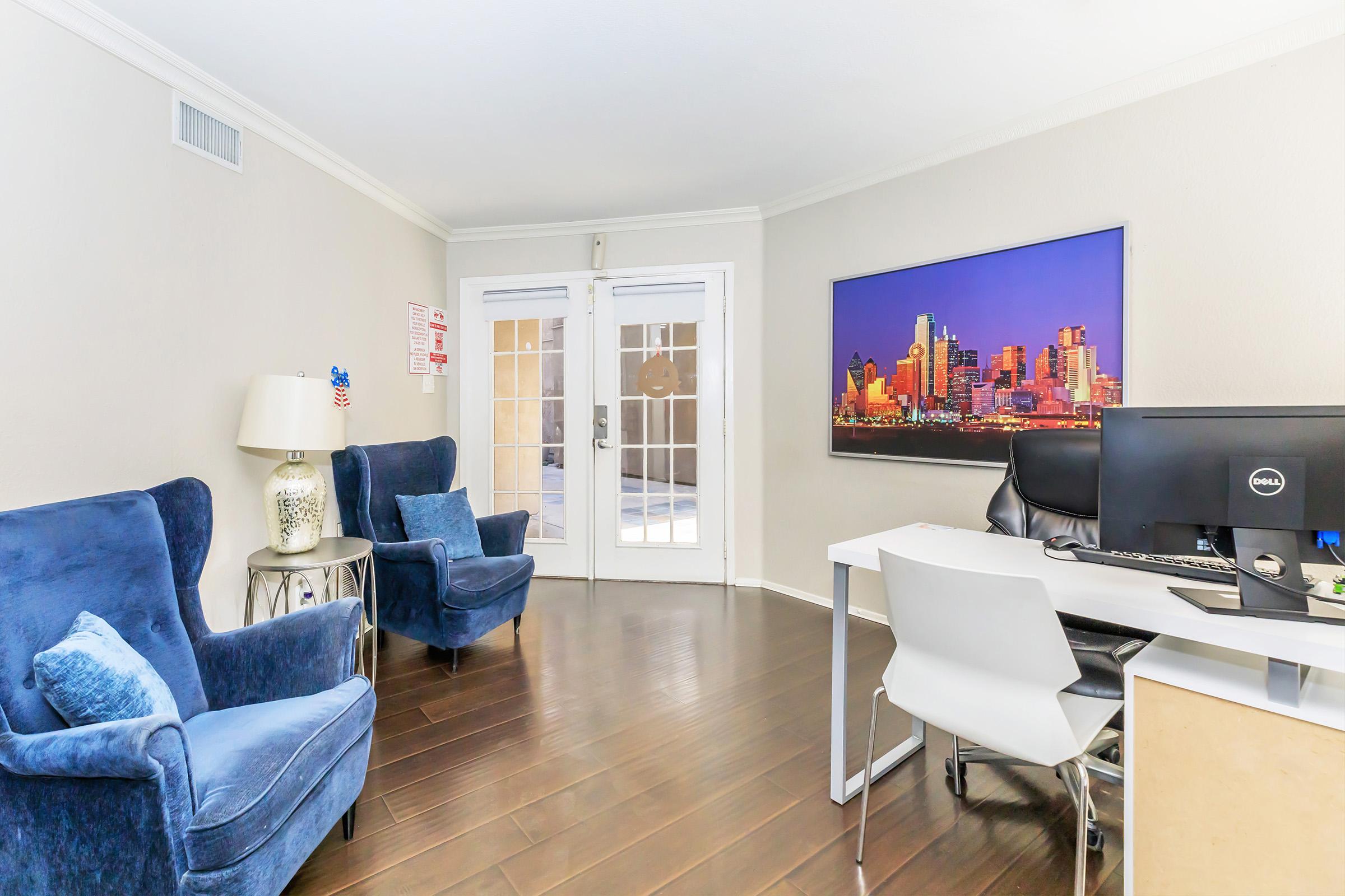 A modern office space featuring two blue armchairs, a desk with a computer, and a large wall-mounted photo of a city skyline. The room has wooden flooring and access to an outdoor area through glass doors.
