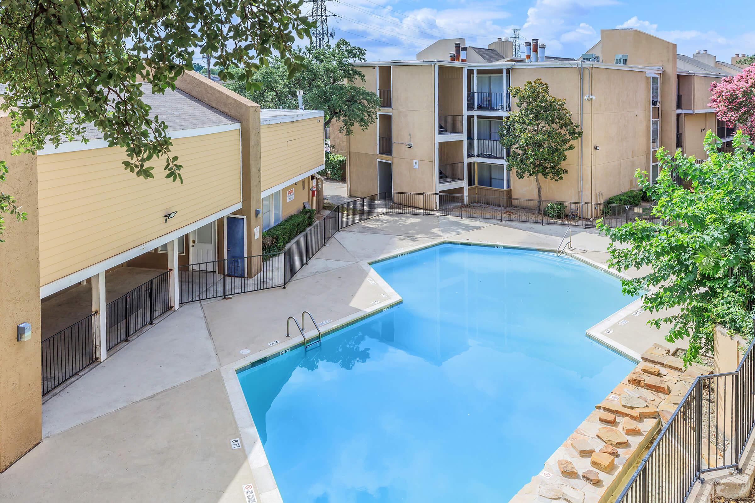 Clear blue swimming pool surrounded by a tan and cream-colored apartment complex. Lush green trees and pink flowering plants provide a touch of nature. The area is well-maintained, featuring a poolside patio with seating. The sky is bright with a few scattered clouds. Ideal for relaxation and leisure activities.